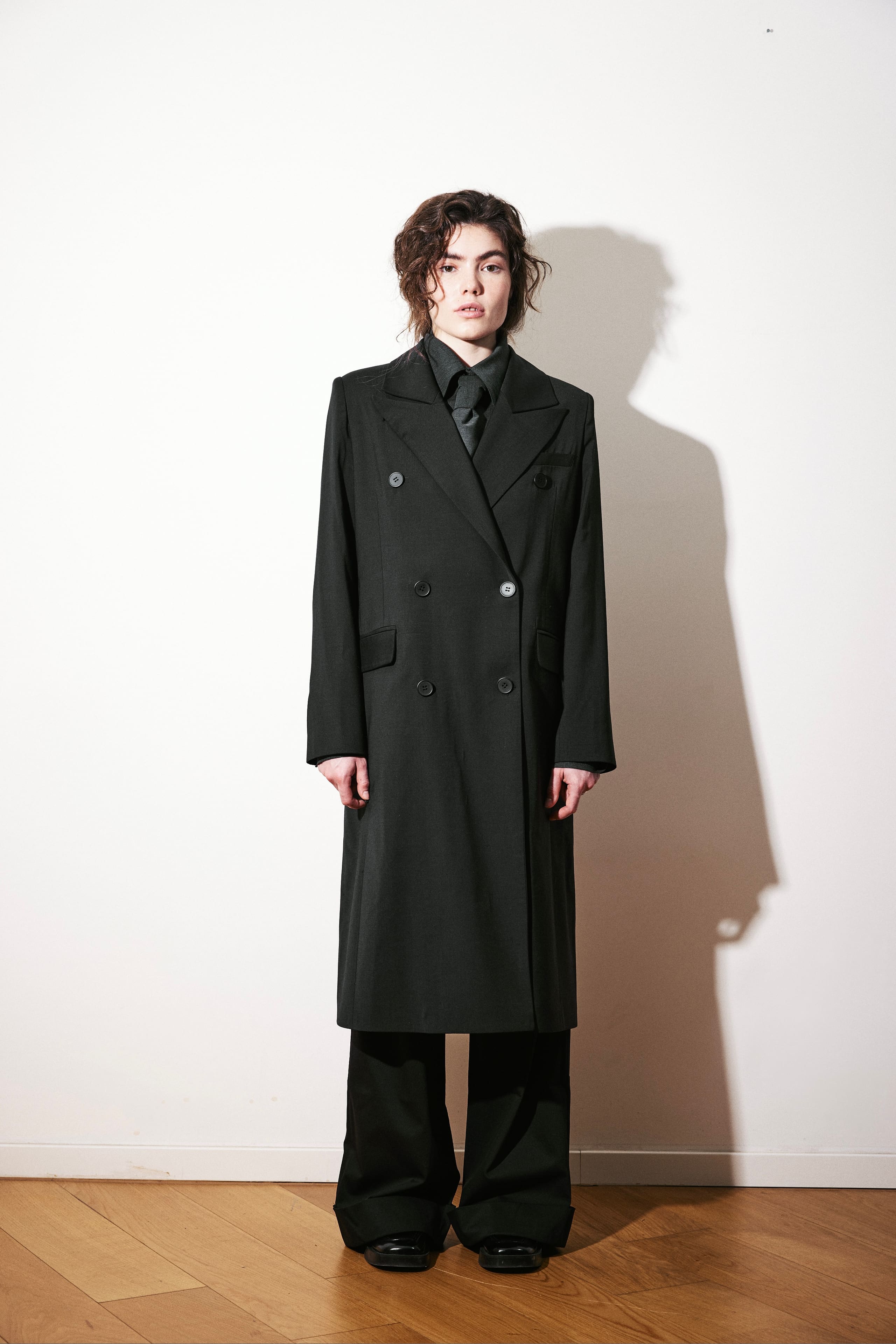 AGIC's black double-breasted overcoat worn full length over a grey shirt and knotted tie.