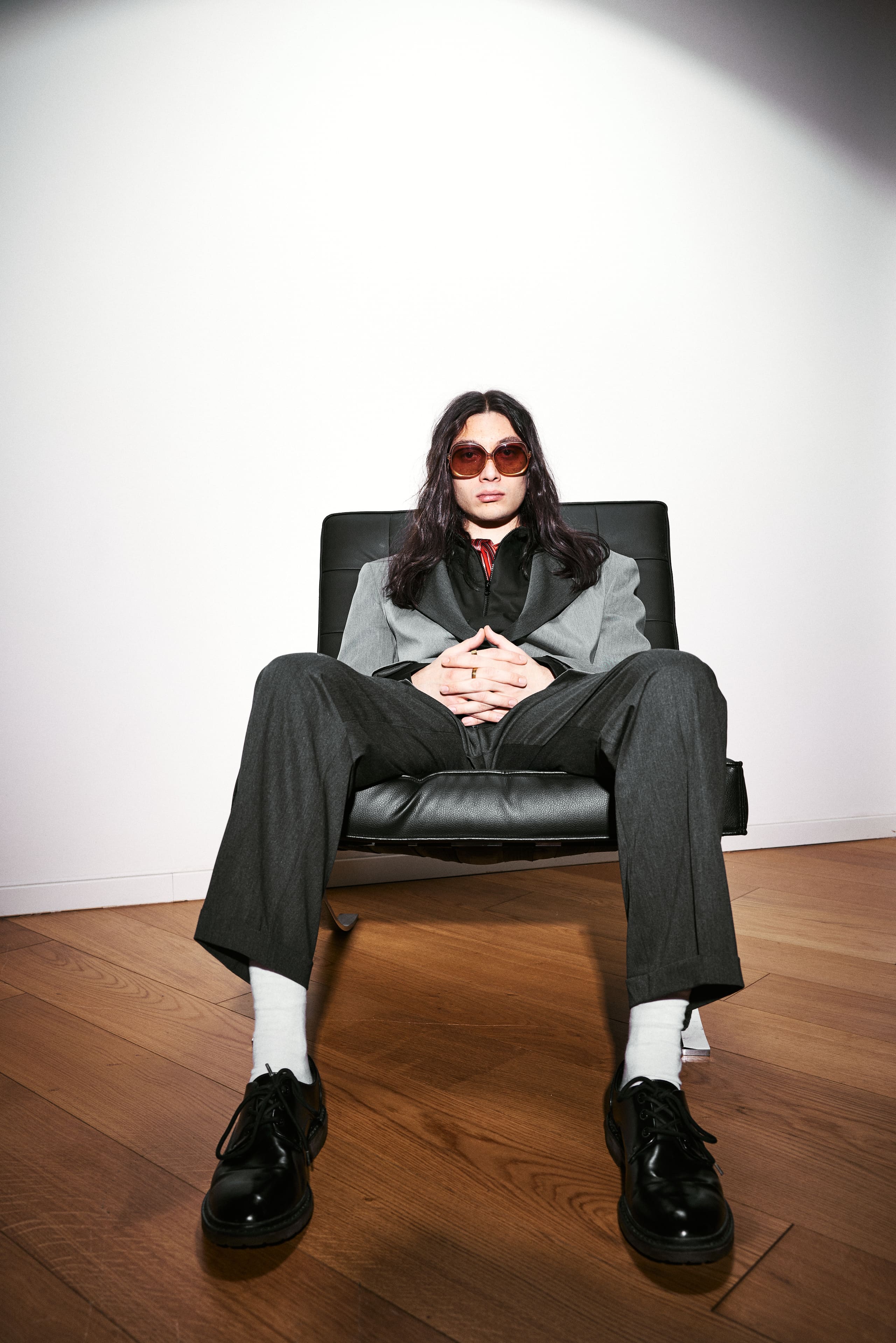 Shot from the floor, a seated frontal pose in AGIC grey tailoring with white socks and polished derbies.
