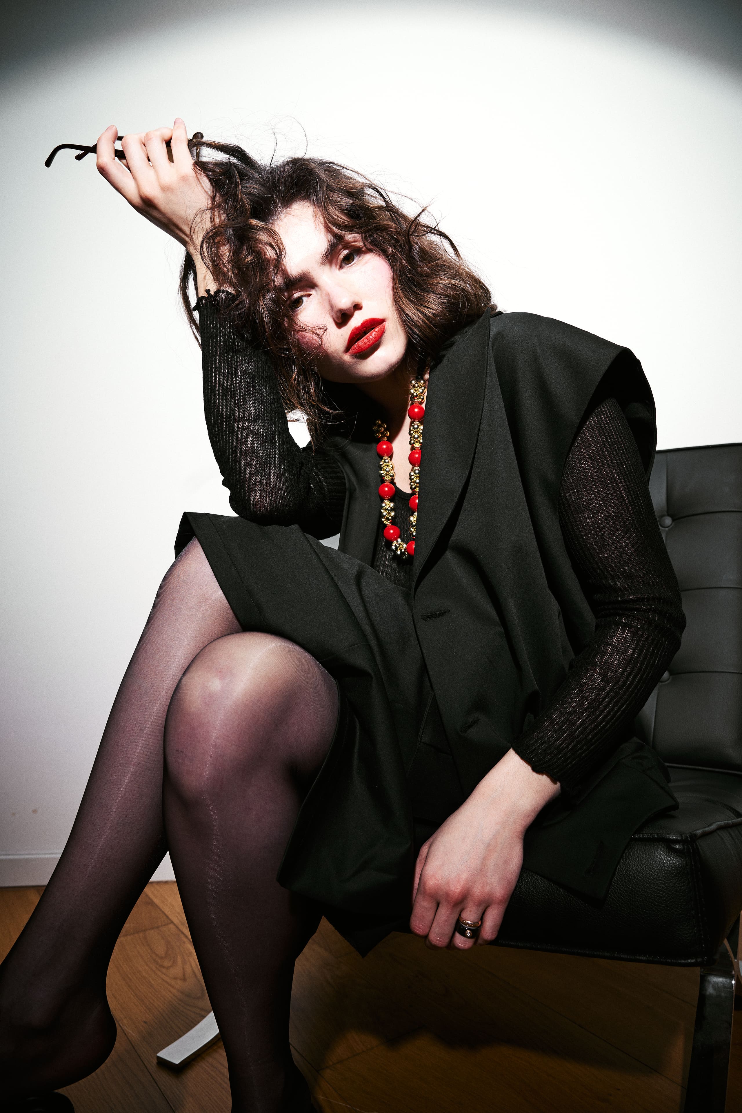 Crossed legs in sheer black and a red bead necklace lift AGIC's shawl-collar vest out of monochrome.