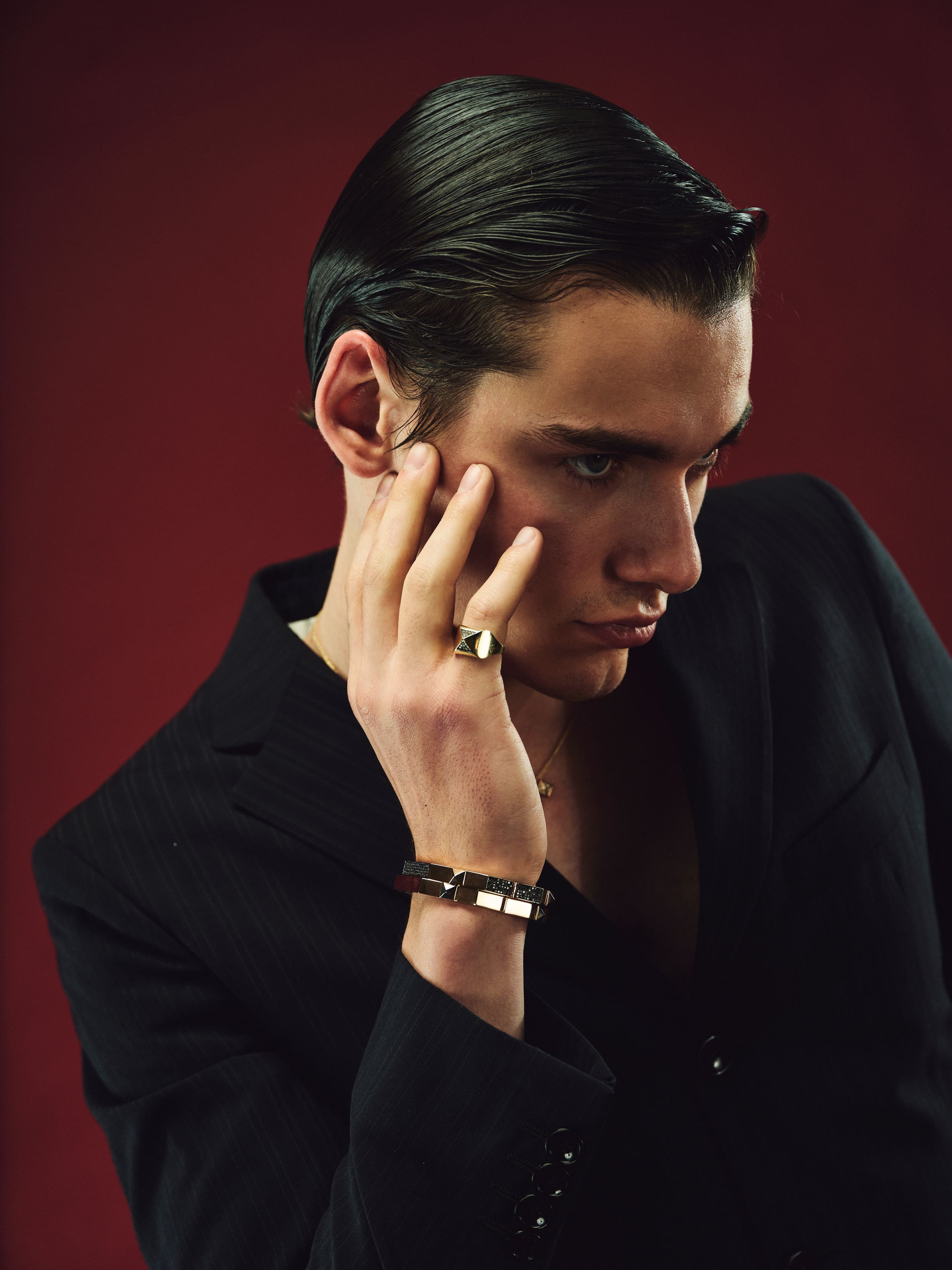 A gold Dime Tribe signet and studded cuff against a slicked profile on deep oxblood red.
