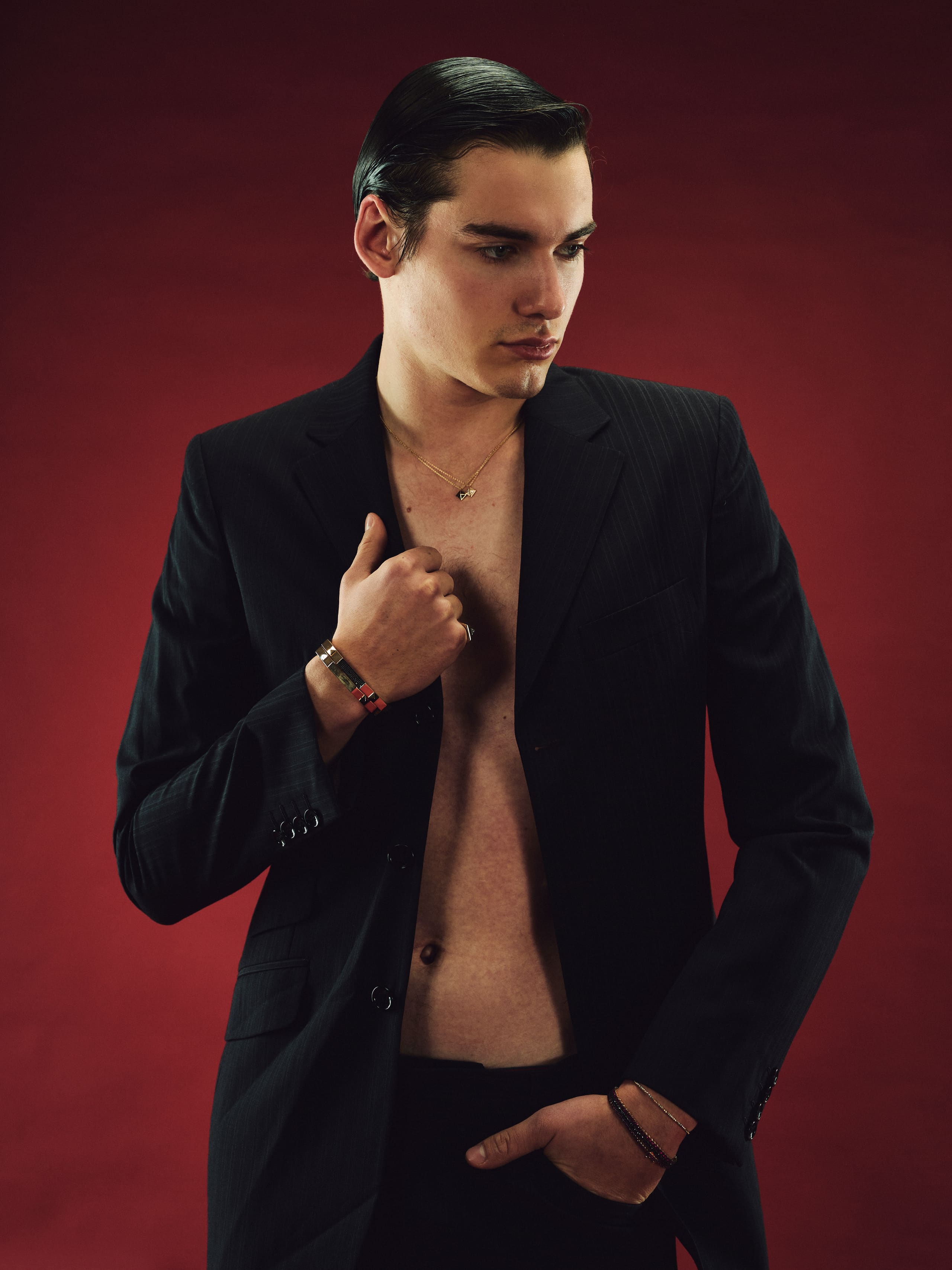 An open pinstripe suit with layered Dime Tribe pendants against a crimson backdrop.