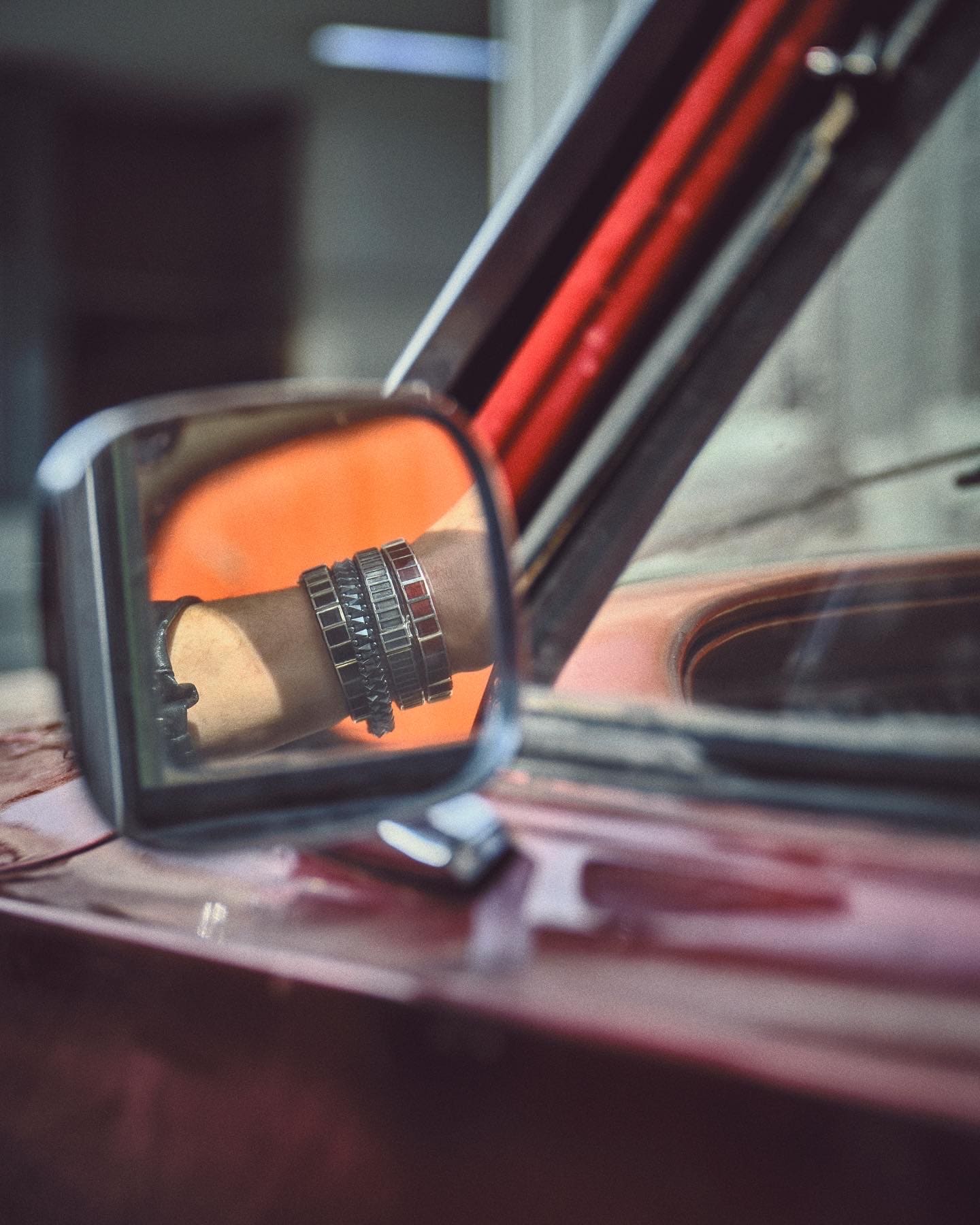 A wing-mirror reflection catches a wrist of stacked Dime Tribe baguette bracelets against the red flank of a classic car.