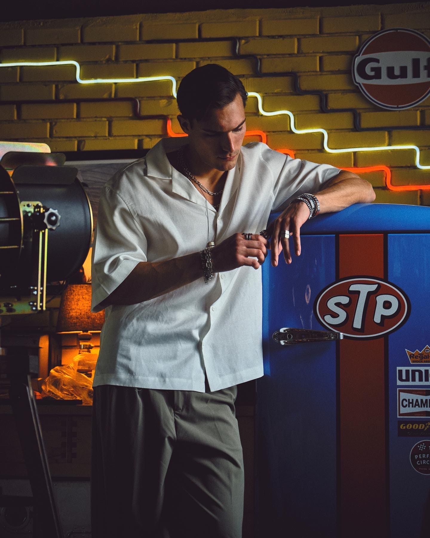 In a white camp-collar shirt and silver Dime Tribe stacks, the model leans on a Gulf-liveried fridge beneath yellow-brick neon.