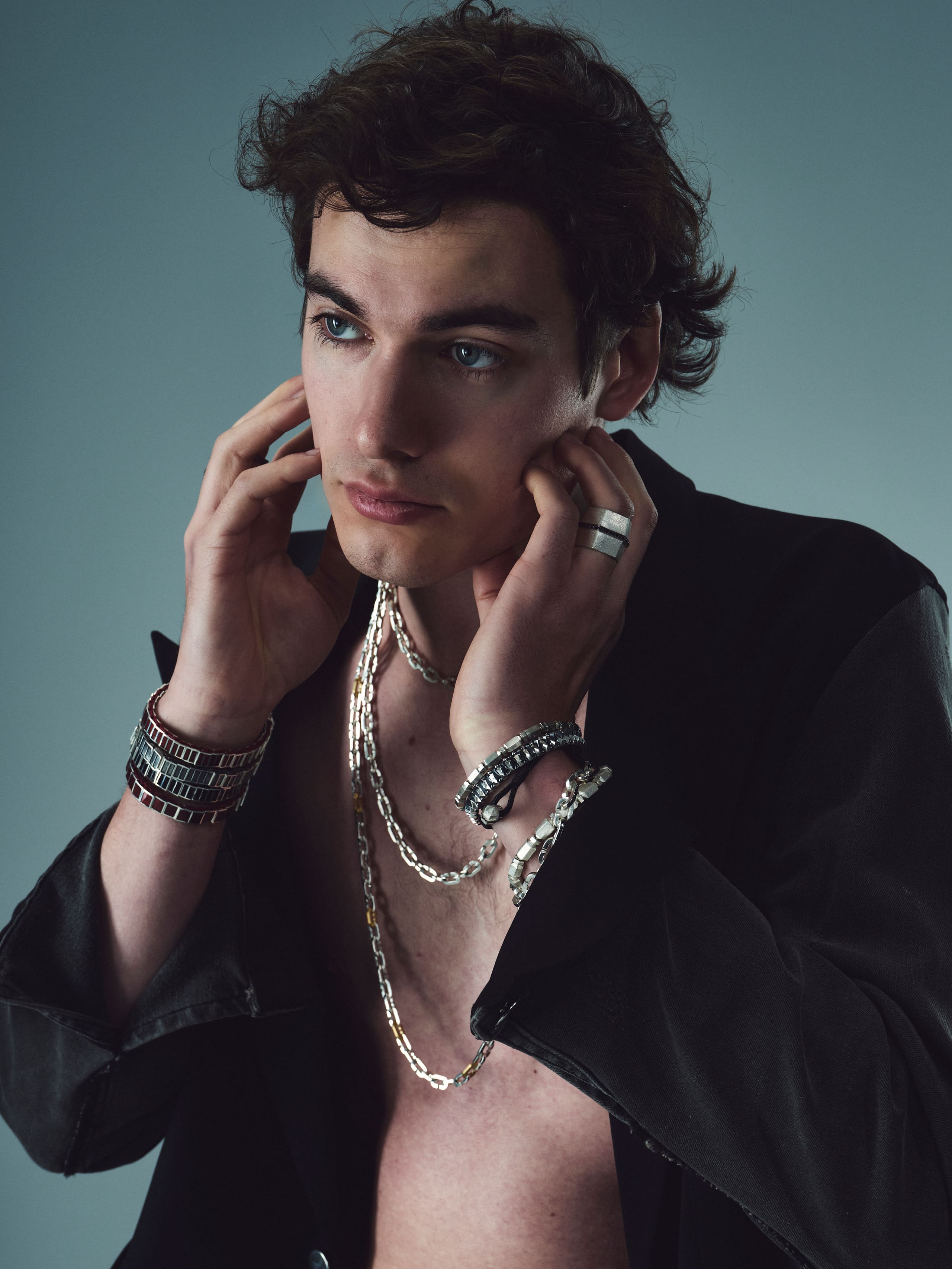 A curly-haired model in an open black jacket layers Dime Tribe chains, cuffs and rings against a slate studio backdrop.