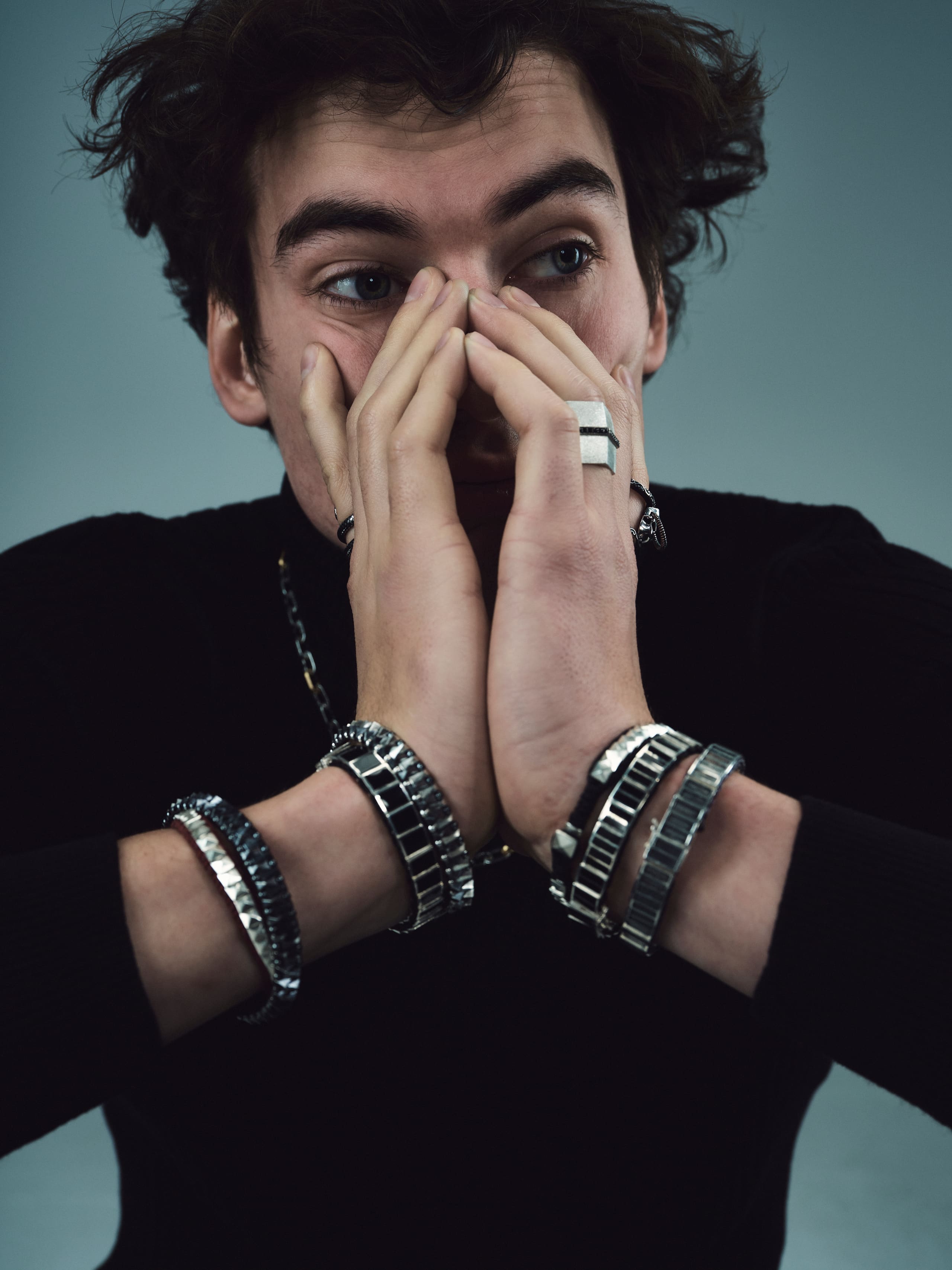 Hands ringed with Dime Tribe bracelet stacks shield a wide-eyed stare in the studio.