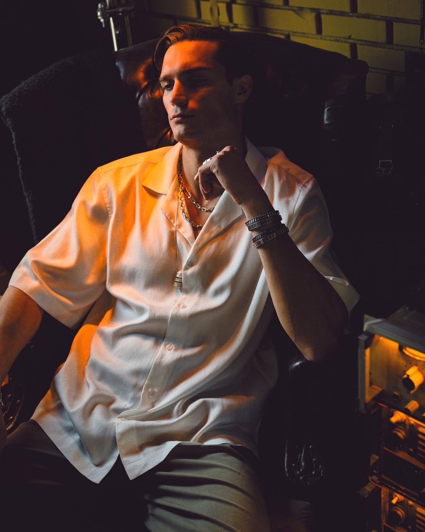Amber rim light skims a satin shirt and Dime Tribe cuffs as the model sinks into a leather armchair.