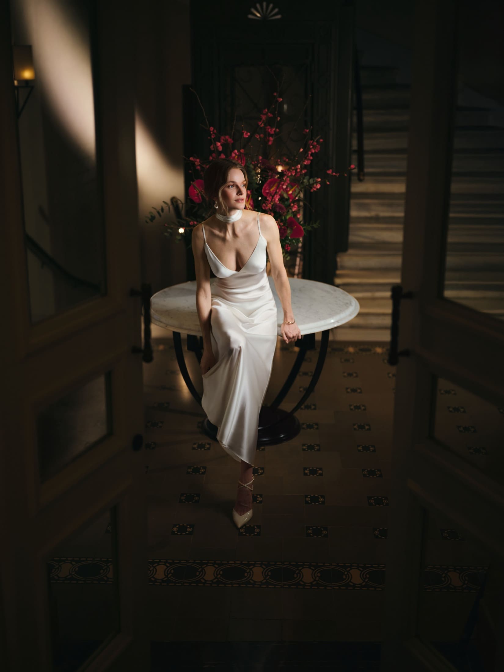 A model in an ivory silk slip dress perches on a marble table in a dim historic foyer for Emelia