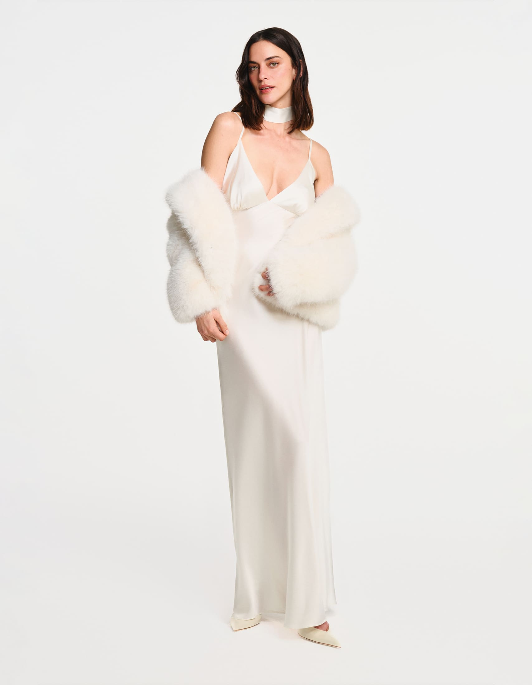 A full-length look at an ivory silk gown worn with a marabou-feather stole against a white studio wall for Emelia