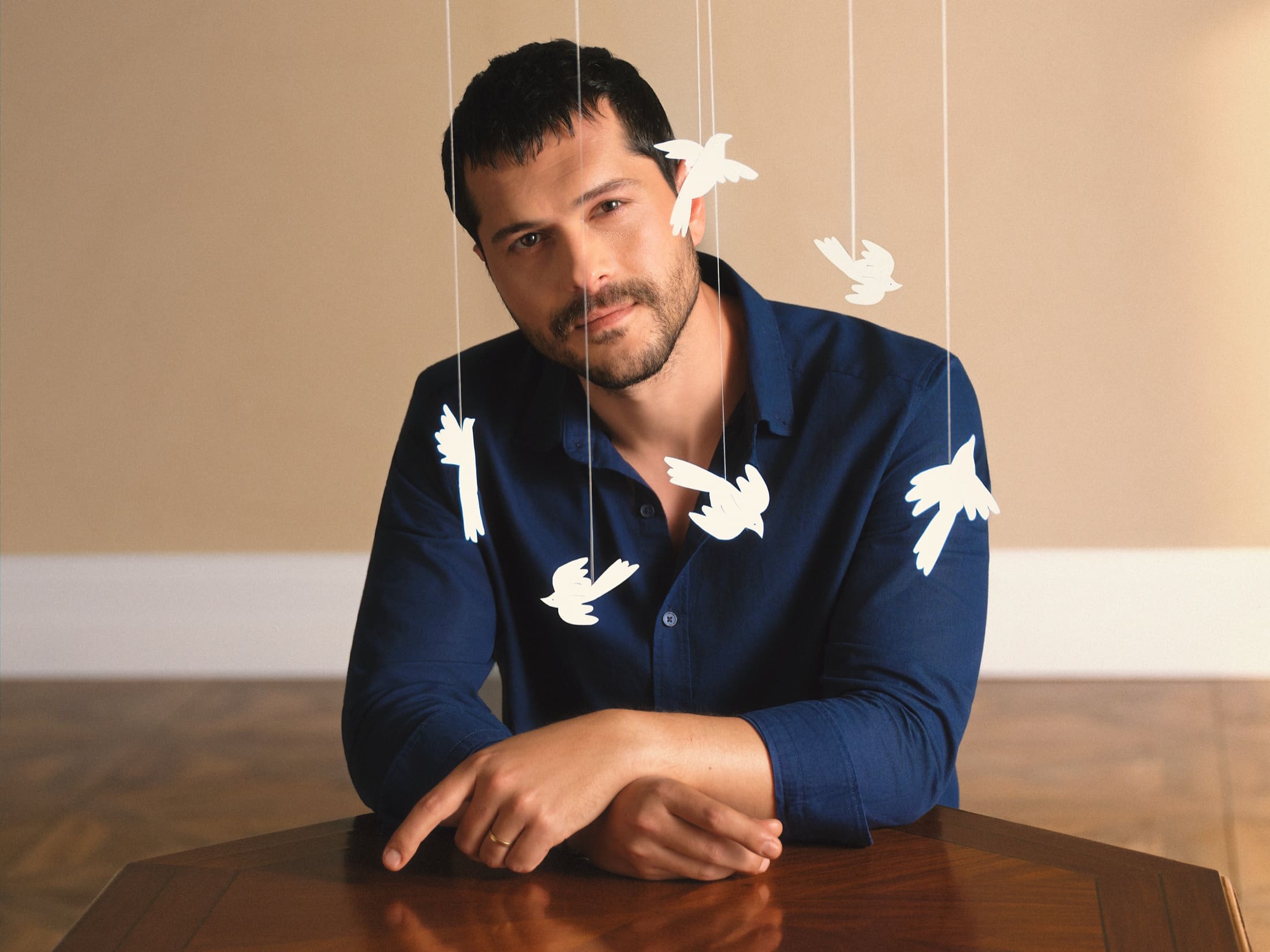 Alperen Duymaz rests his arms on a polished wooden table in a navy shirt, ringed by paper doves suspended on threads, for LC Waikiki's Father's Day campaign.