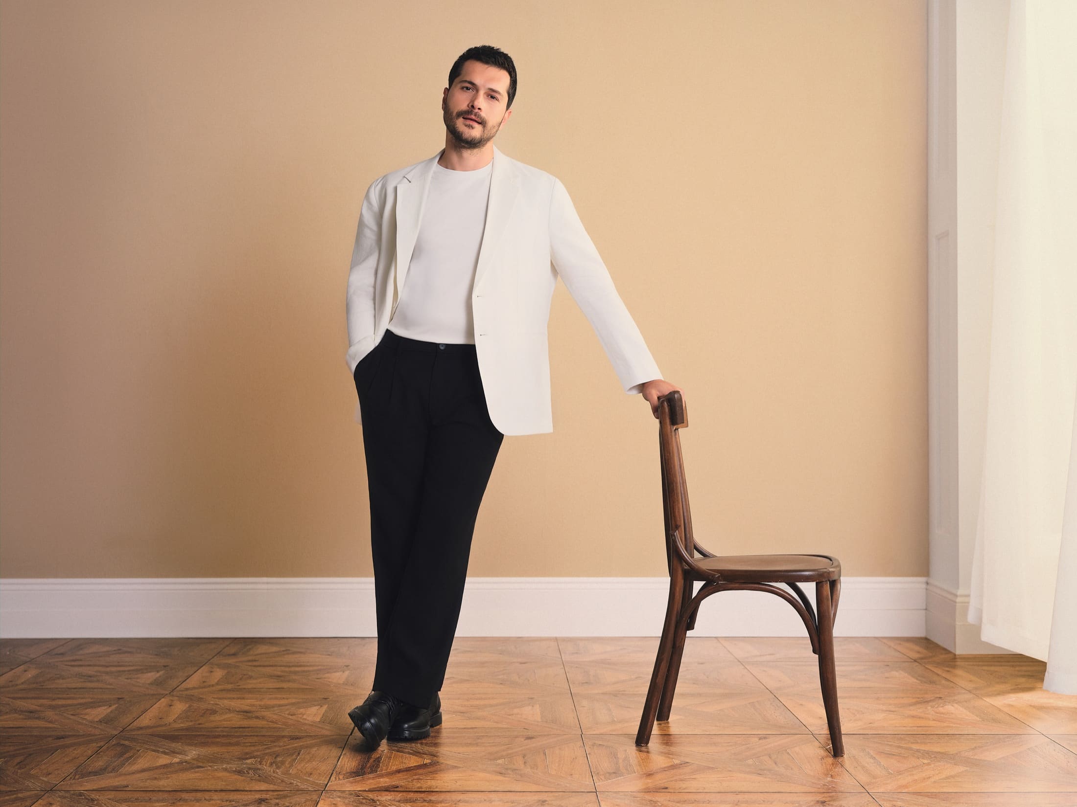 Alperen Duymaz stands full-length on parquet in an off-white blazer and black pleated trousers, one hand resting on a bentwood chair, for LC Waikiki.