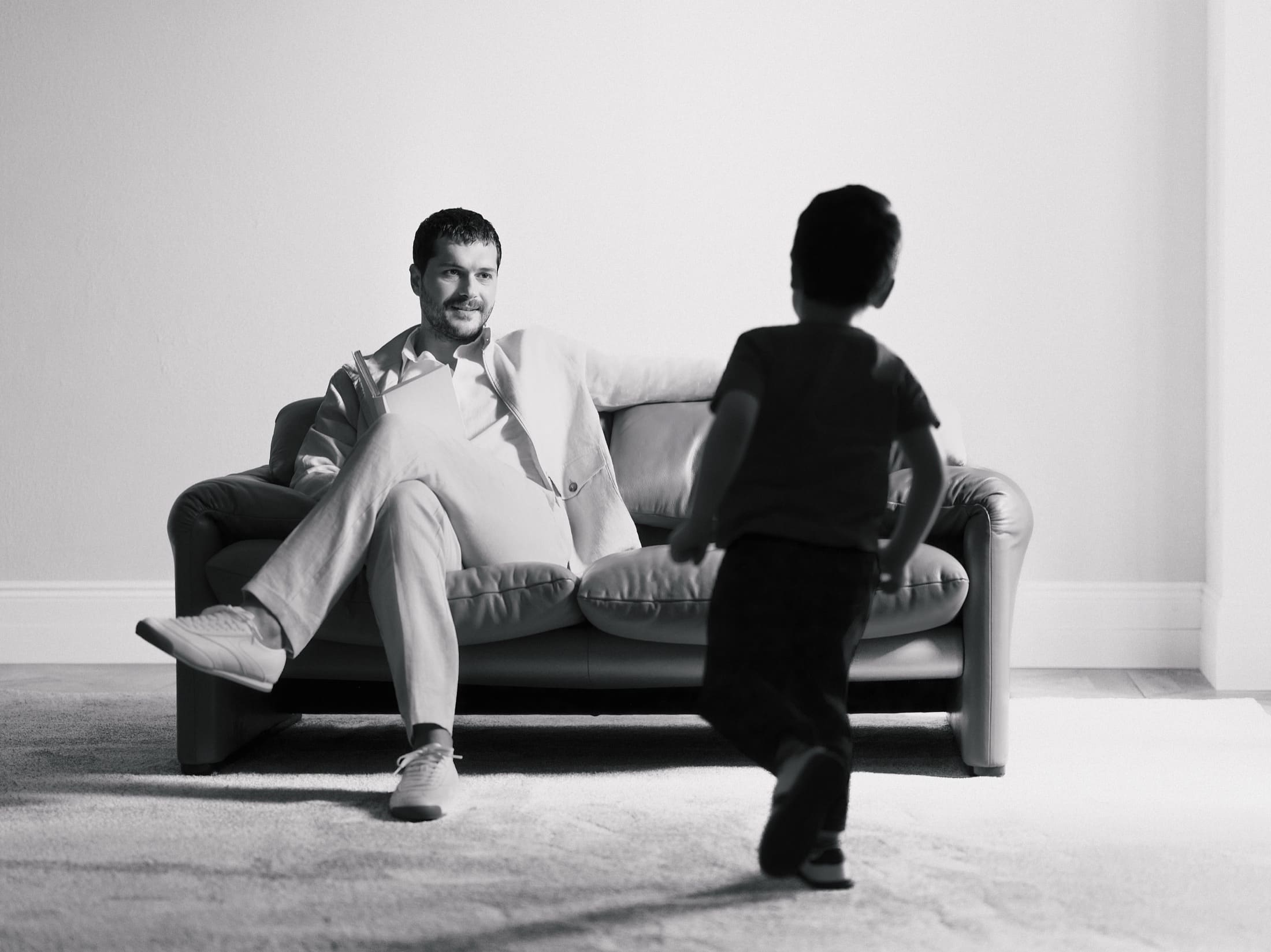 In black and white, Alperen Duymaz looks up from his book on a leather sofa as a young boy runs toward him, closing LC Waikiki's Father's Day story.