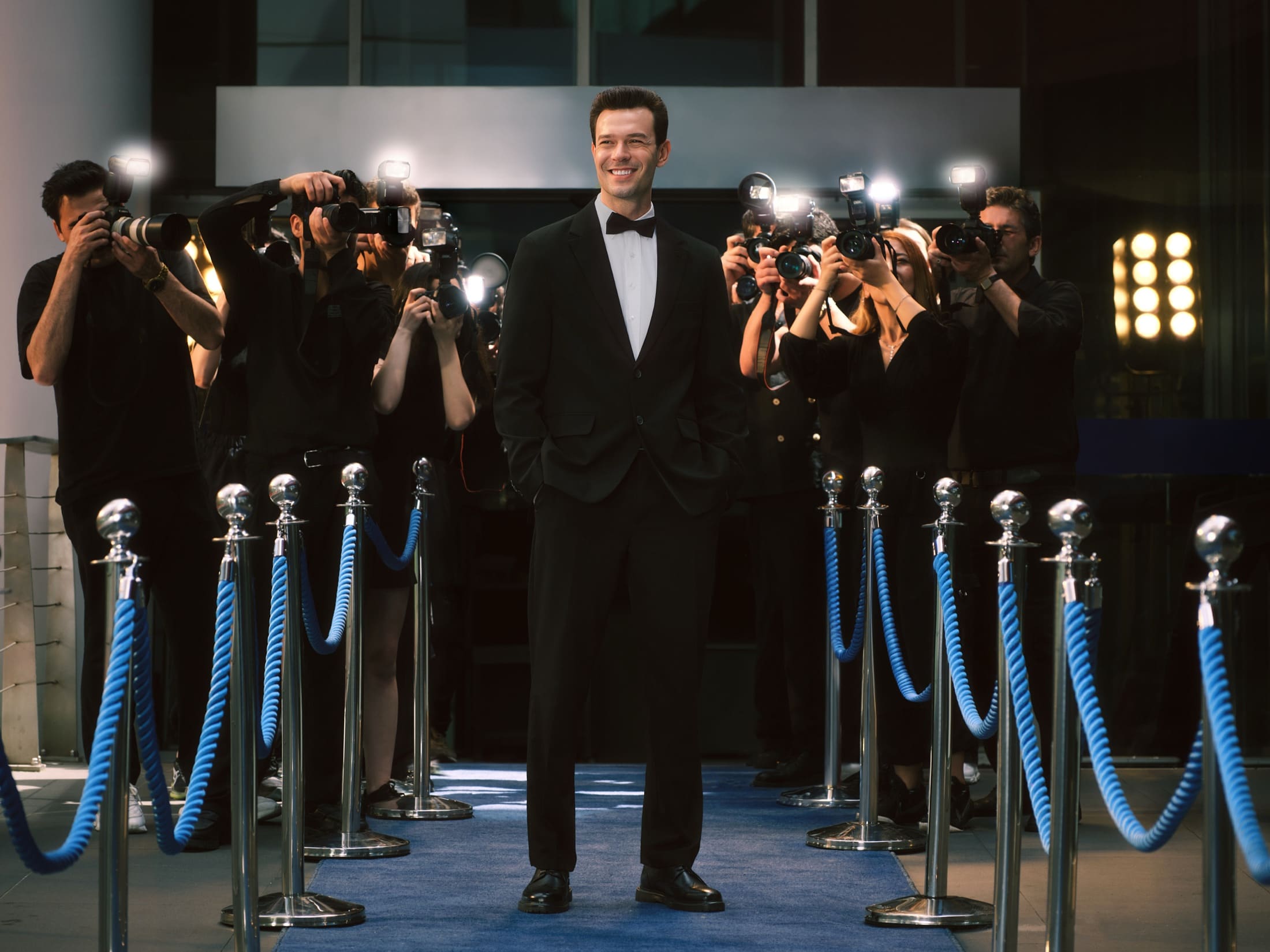 Aytaç Şaşmaz stands in a black tuxedo on a blue carpet, hands in pockets and grinning as a wall of paparazzi flashes goes off behind the ropes, for LC Waikiki.