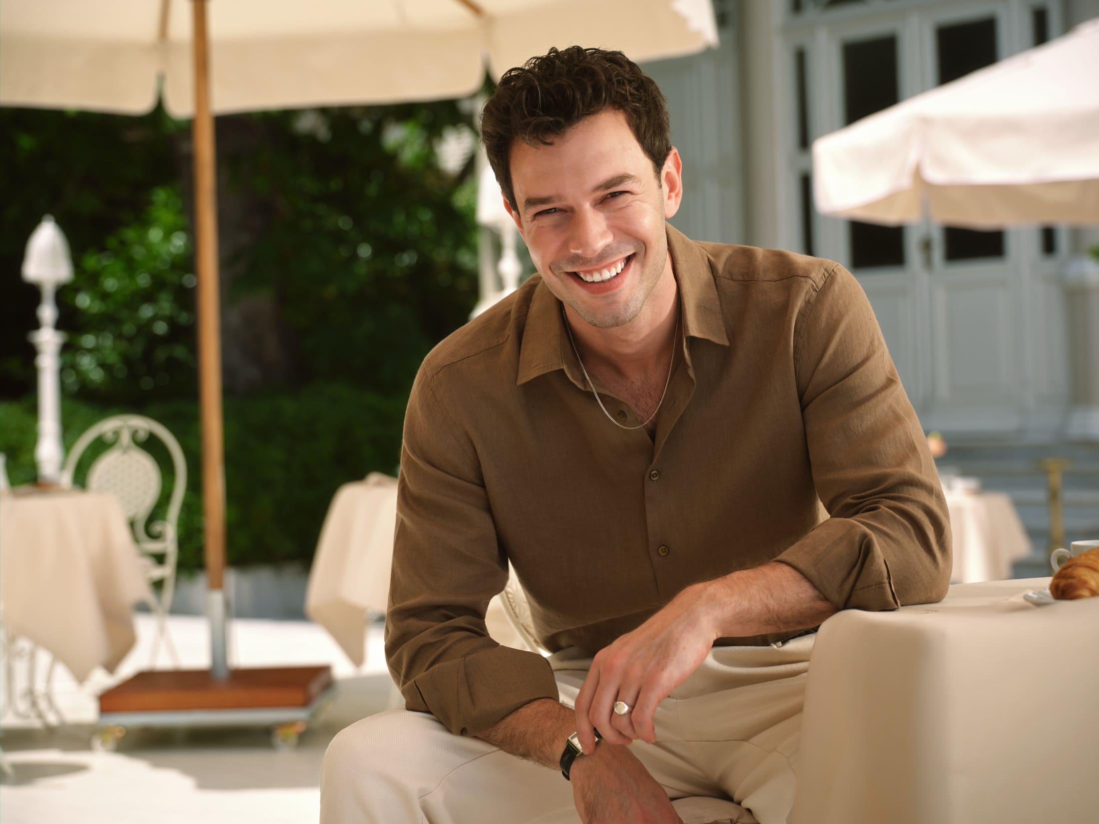 A relaxed close-up of Aytaç Şaşmaz laughing on a sunlit terrace in an open brown linen shirt and slim silver chain for LC Waikiki.