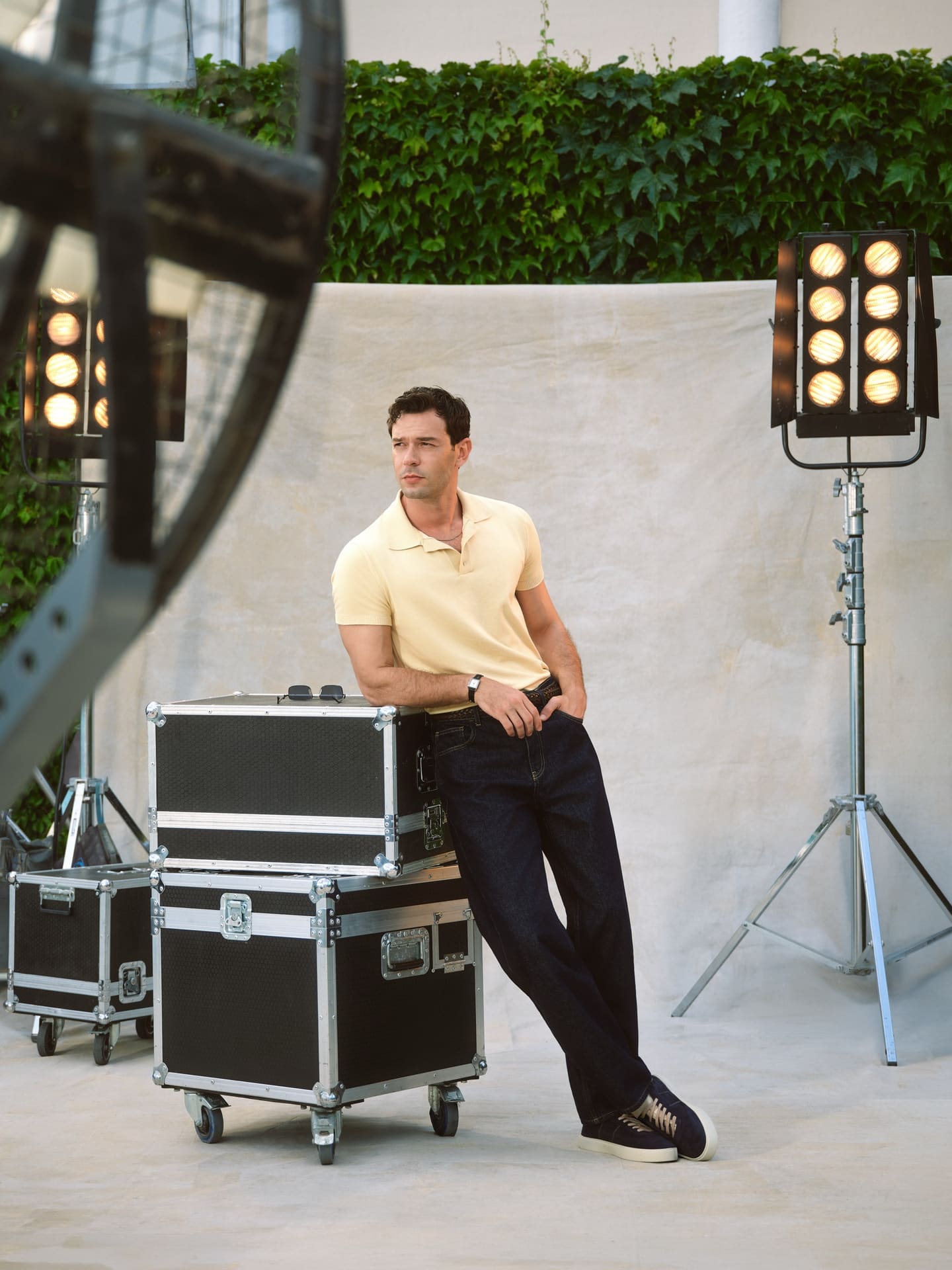 Between takes, Aytaç Şaşmaz leans on stacked flight cases in a pale yellow knit polo and dark selvedge jeans, framed by tungsten heads on a canvas backdrop, for LC Waikiki.