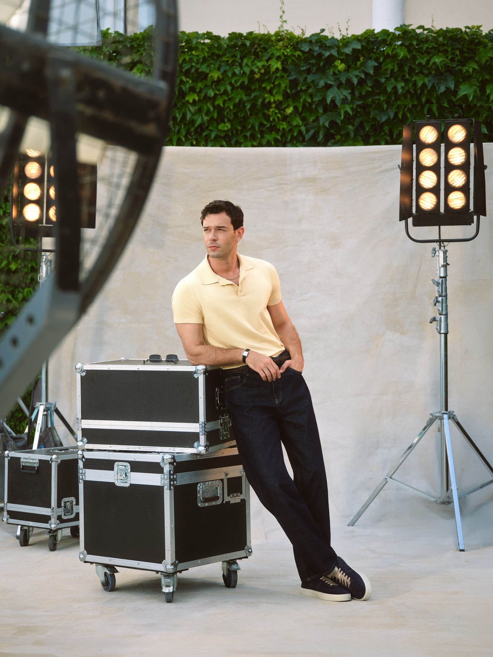 Between takes, Aytaç Şaşmaz leans on stacked flight cases in a pale yellow knit polo and dark selvedge jeans, framed by tungsten heads on a canvas backdrop, for LC Waikiki.