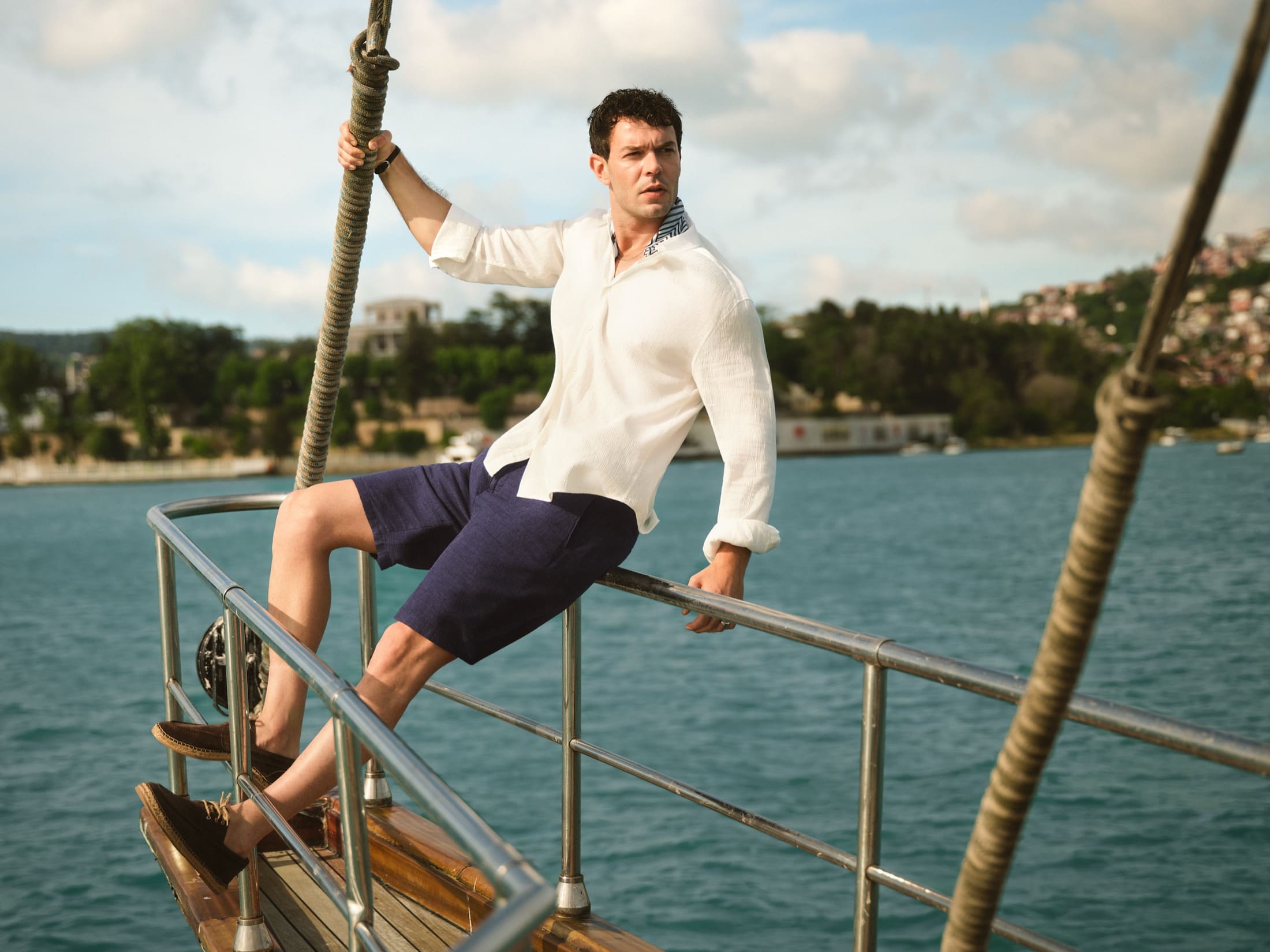 Aytaç Şaşmaz perches on the chrome rail of a Bosphorus gulet, one hand on the rigging, in a crinkled white shirt and navy linen shorts for LC Waikiki.