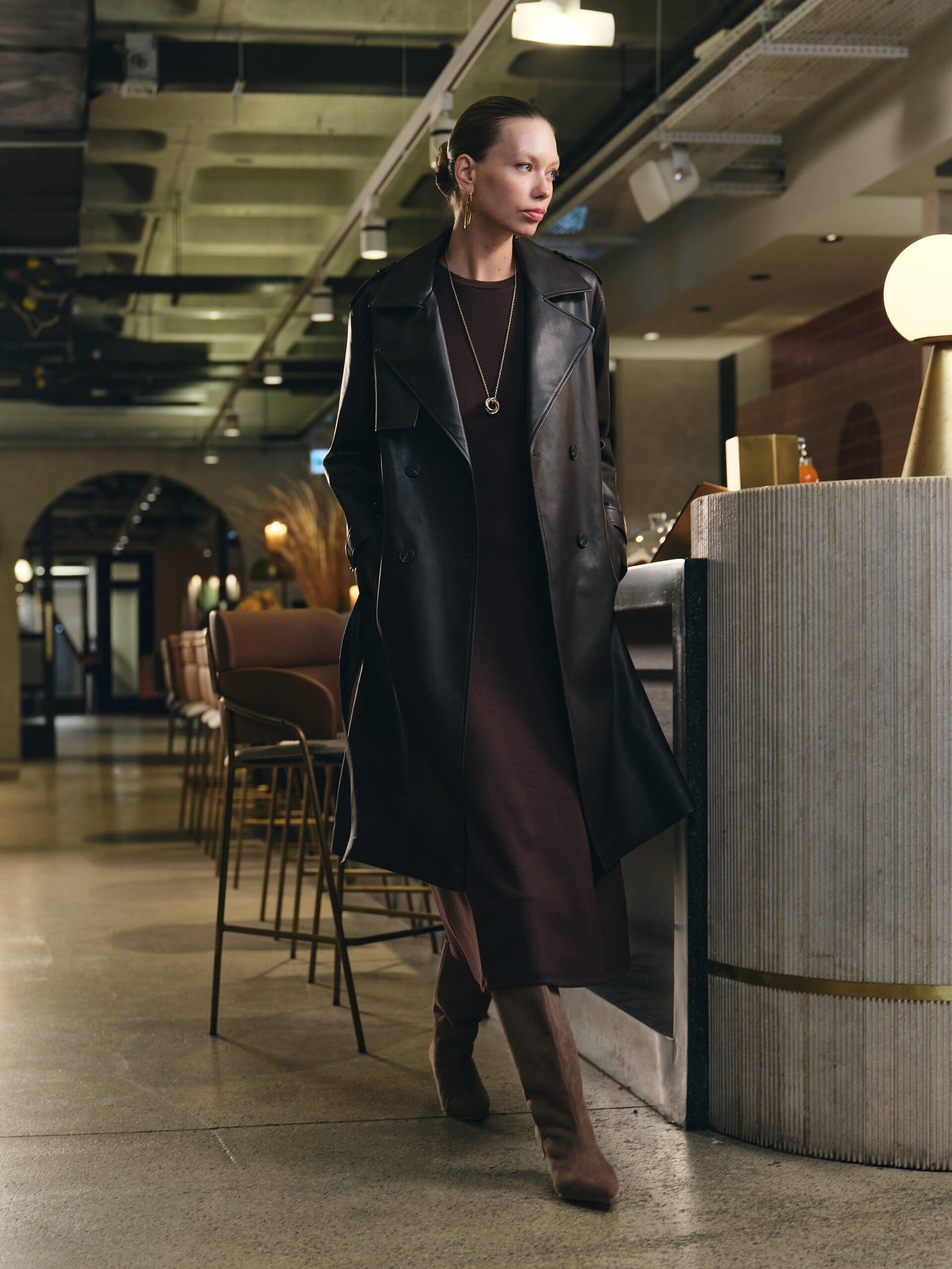 The black leather trench worn over a chocolate midi dress, paused mid-stride along the bar's fluted counter for LC Waikiki