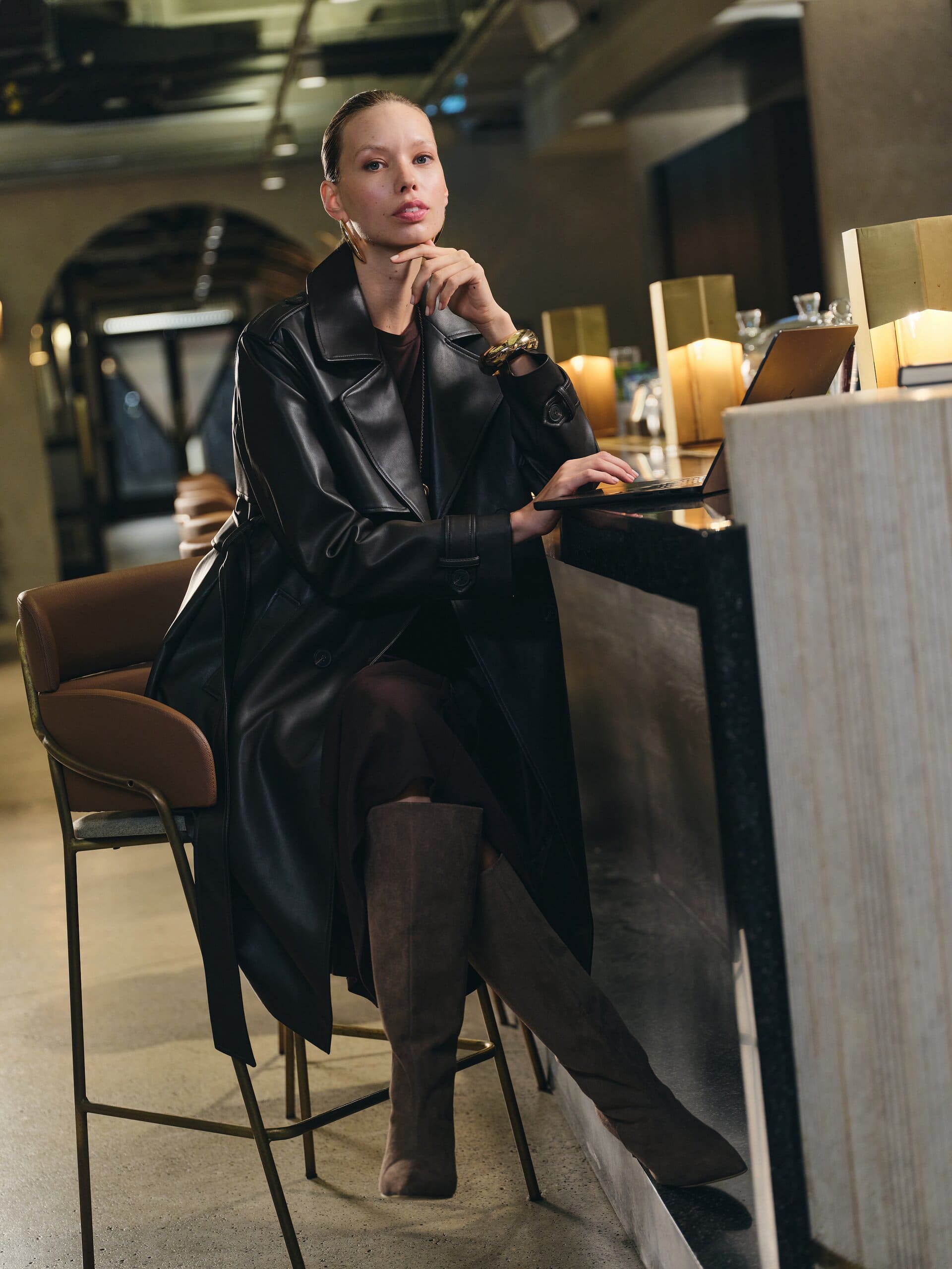 A model in a black leather trench and suede knee boots works a laptop at a dim bar counter for LC Waikiki