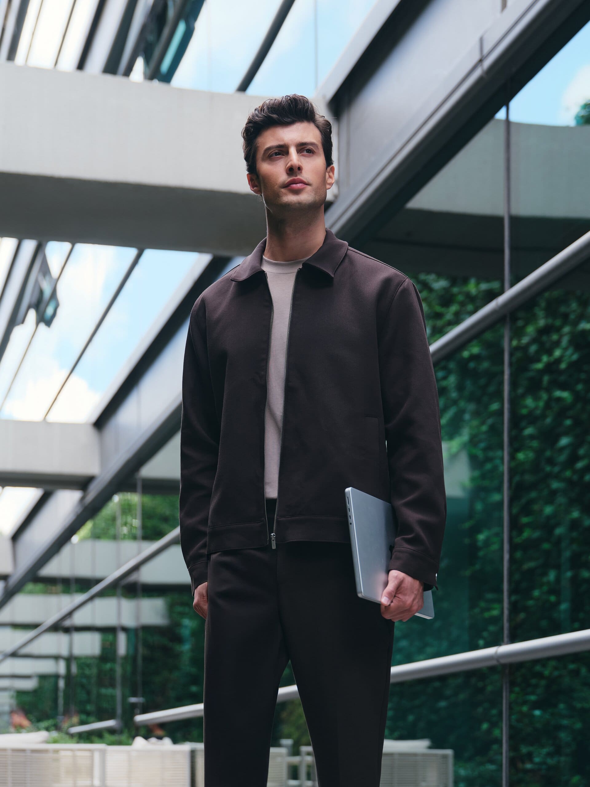 Shot from below against a glass atrium, a model in a dark brown zip jacket carries his laptop like a briefcase for LC Waikiki