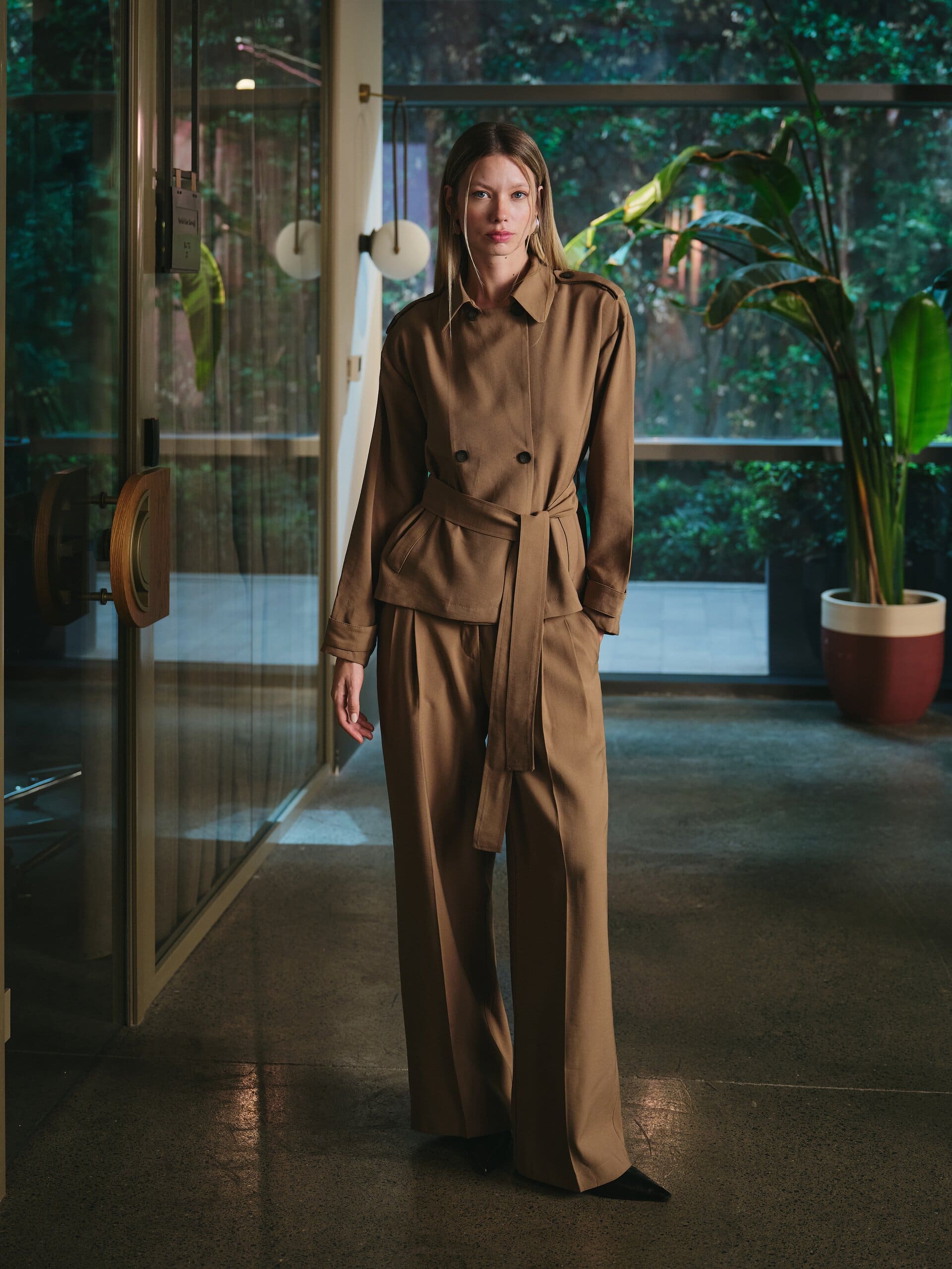 Head-to-toe tobacco tailoring, a belted double-breasted jacket over pooling wide-leg trousers, squared to the camera in a glass corridor for LC Waikiki