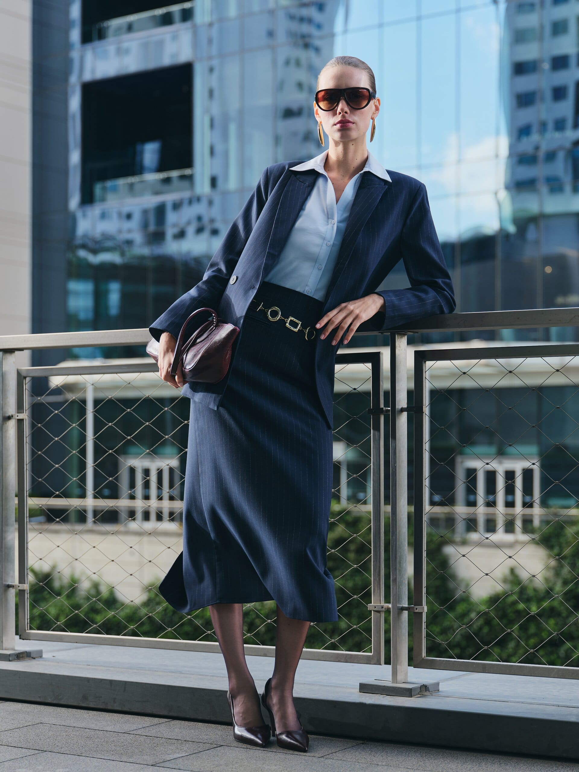 In aviator shades and a navy pinstripe skirt suit, a model leans on a mesh railing before mirrored towers for LC Waikiki