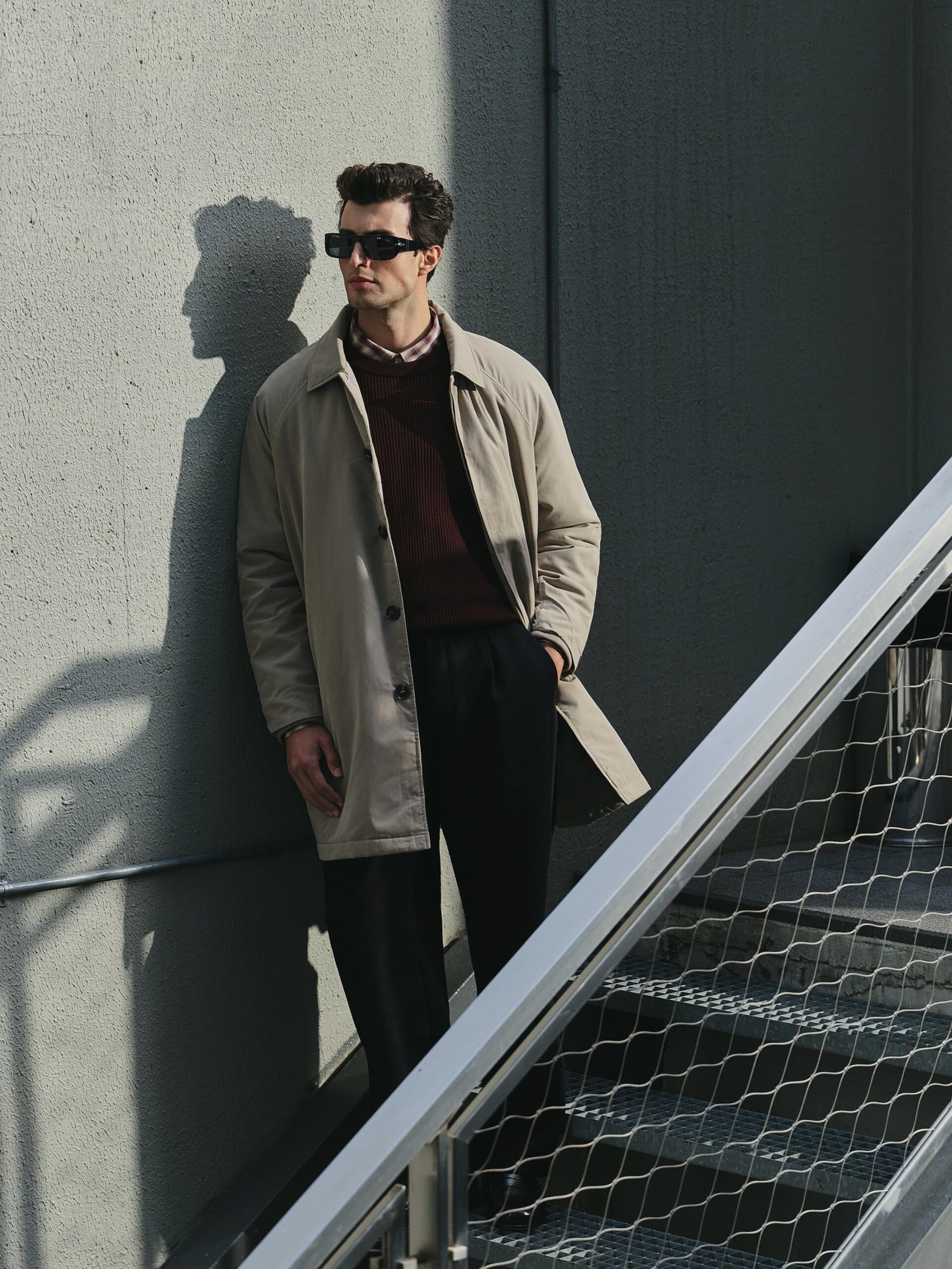 The stone mac throws a long shadow on the grey stair wall, hand pocketed beside the steel banister for LC Waikiki