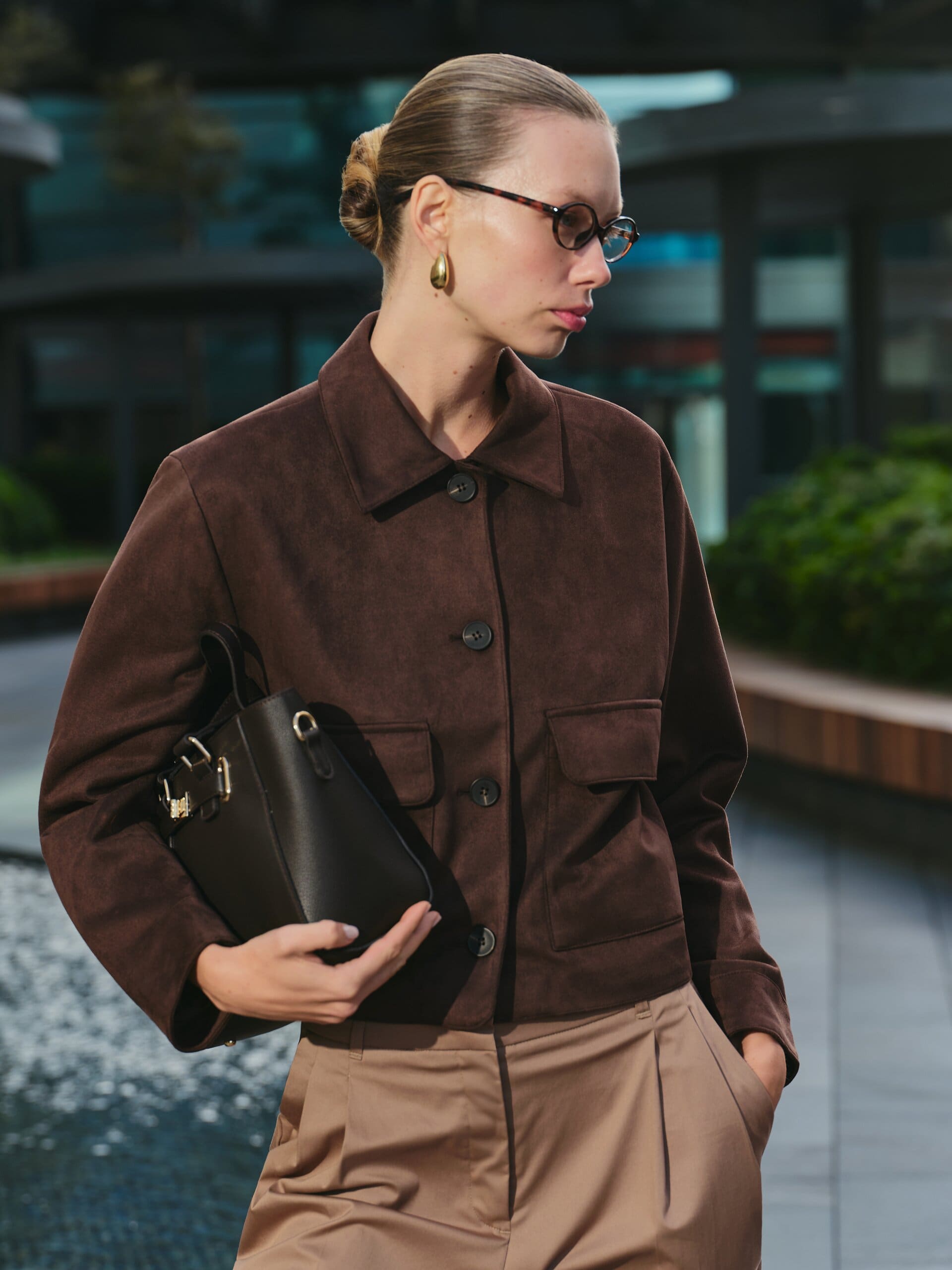 Close over the shoulder in the suede jacket, tortoise glasses turned to the plaza's evening glass for LC Waikiki