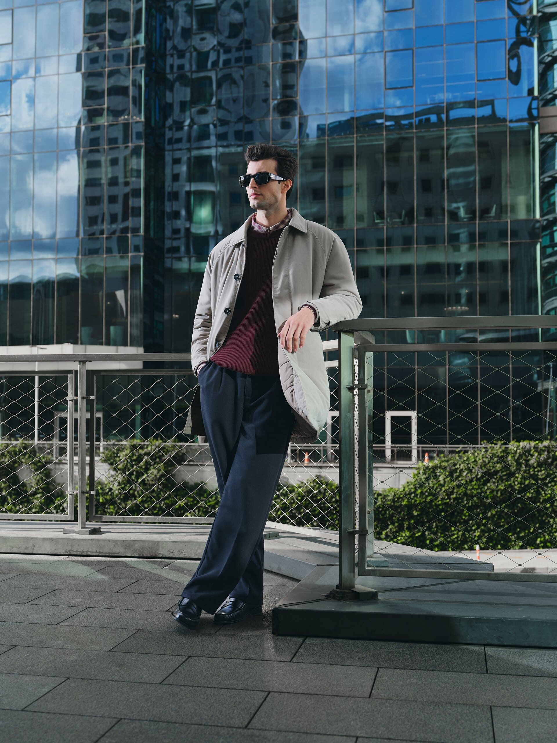 A model in a stone mac and burgundy ribbed knit rests against a walkway railing beneath reflective office glass for LC Waikiki