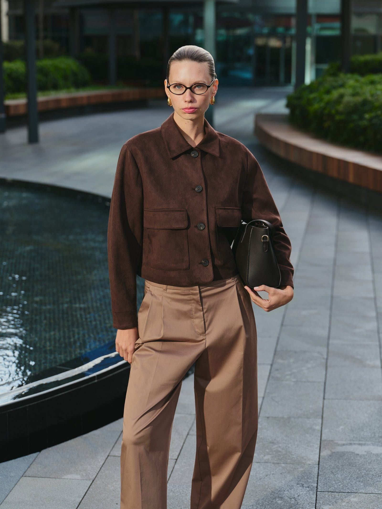 Oval glasses and a chocolate suede jacket with camel trousers hold a steady gaze beside a plaza fountain for LC Waikiki