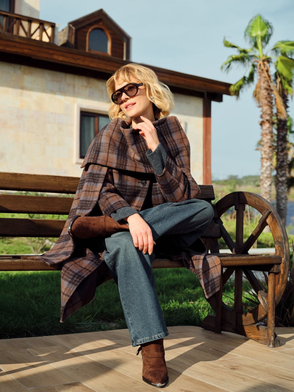 Wrapped in a brown plaid cape coat and oversized sunglasses, Hazal Türesan folds onto a sunlit wooden bench for LC Waikiki's countryside chapter.