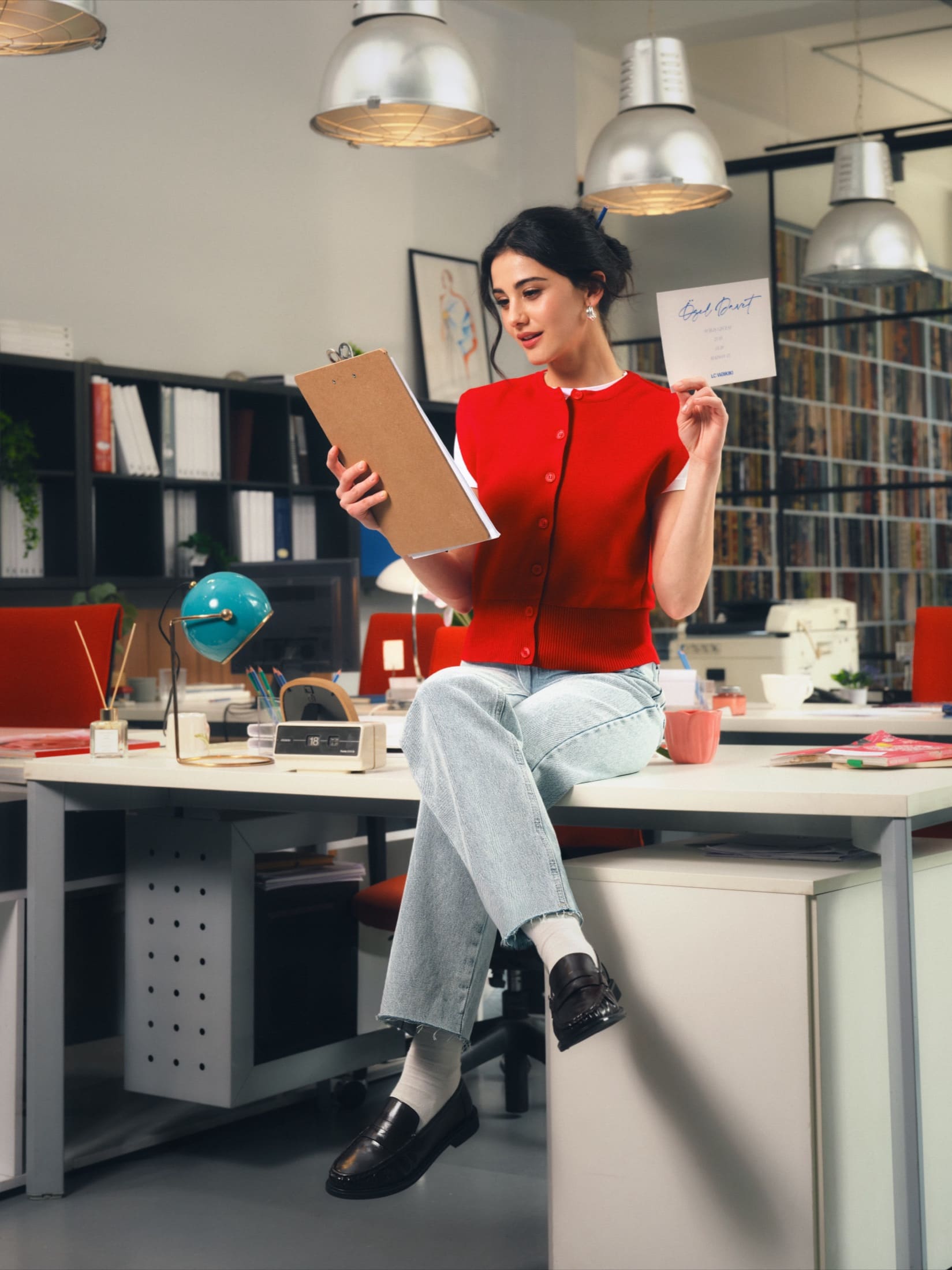 Perched on a desk in a retro design studio, Helin Kandemir reads from a clipboard in a red cropped cardigan and light-wash jeans, holding up an LC Waikiki invitation card.