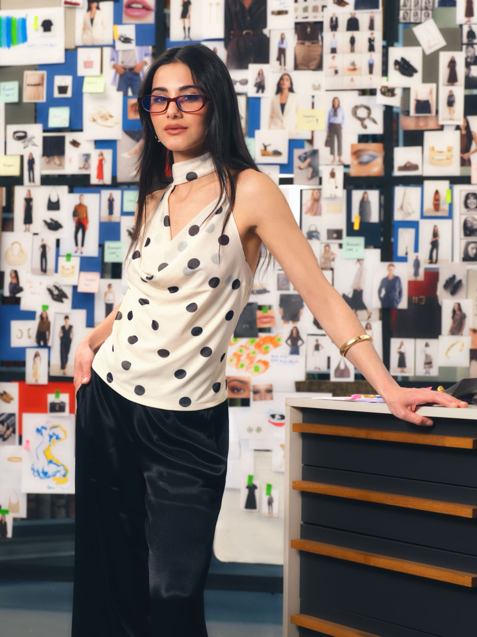 Helin Kandemir poses against a wall of pinned lookbook tears in tinted rectangular glasses, a polka-dot cowl halter and black satin trousers for LC Waikiki's Glow Up story.