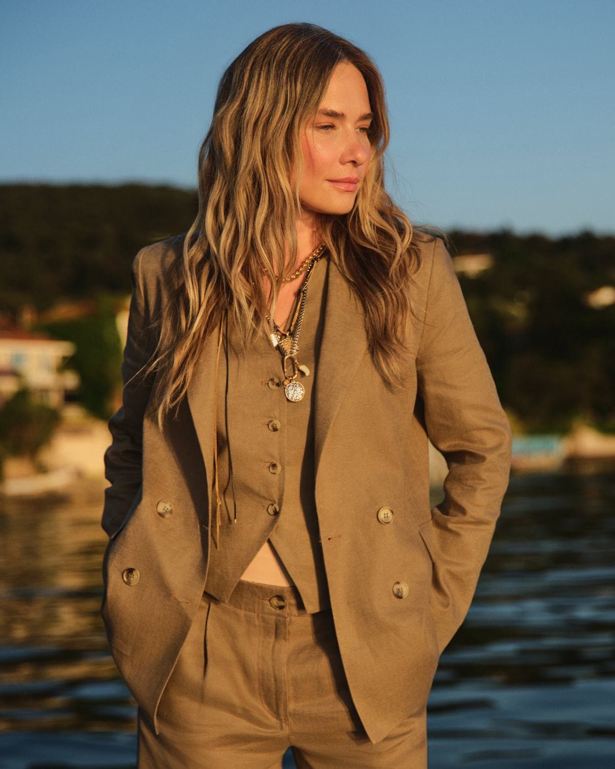 Müge Boz in a taupe double-breasted linen suit layered with coin necklaces, caught in profile at golden hour on the waterline for LC Waikiki