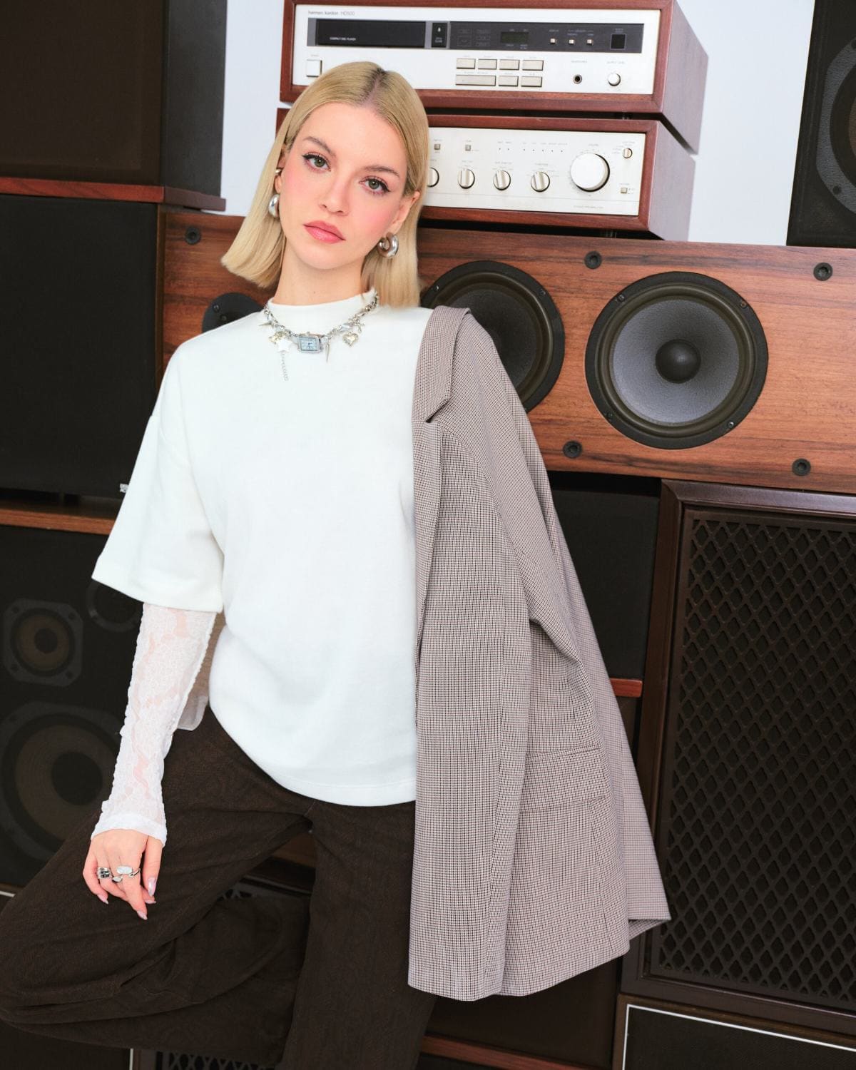Tuana Yılmaz stands before a wall of vintage hi-fi speakers, houndstooth blazer draped over an oversized white tee and lace sleeves for LC Waikiki.