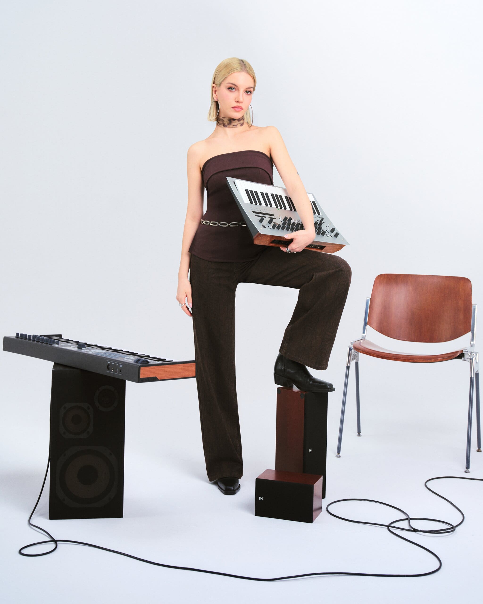 Tuana Yılmaz cradles a vintage synthesizer with one boot on a speaker cab, strapless burgundy top and chain belt in a cabled studio set for LC Waikiki.