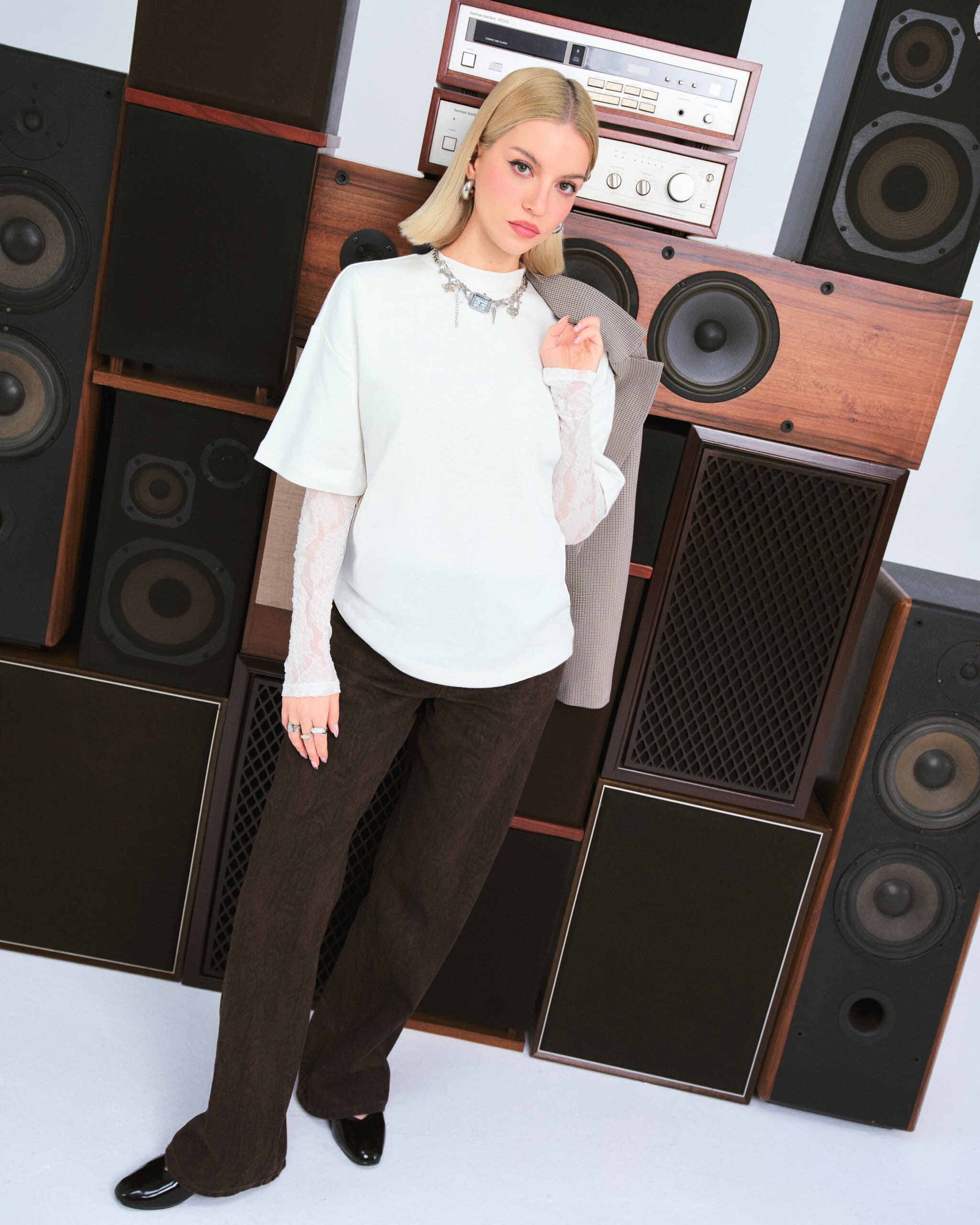 Tilted full-length of Tuana Yılmaz against stacked wood-cased speakers, blazer slung over her shoulder and brown flares grazing the floor for LC Waikiki.