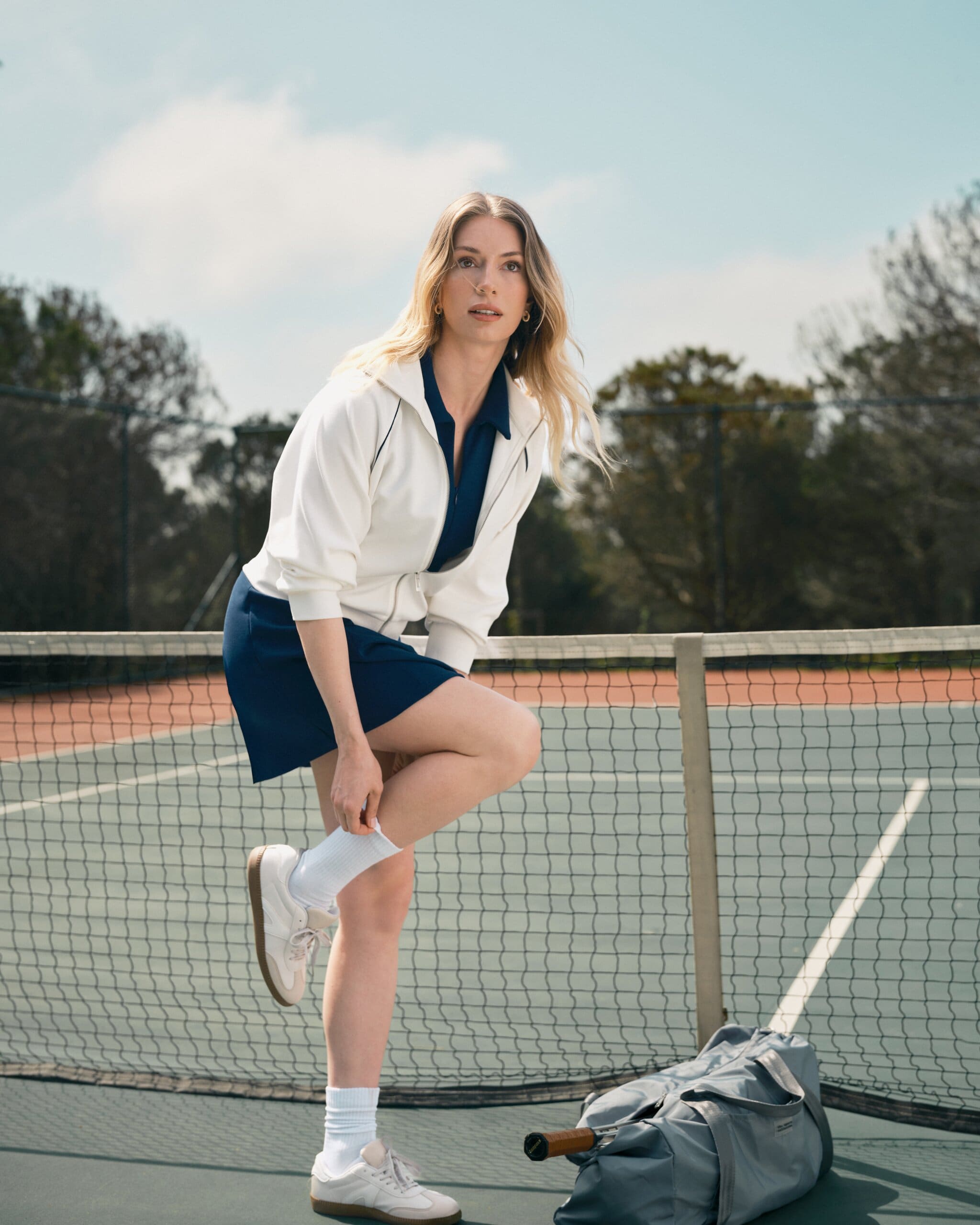 Yasemin Kay Allen crouches at the net to pull up a sock, track jacket over a navy tennis dress, racket and duffel waiting on the court for LC Waikiki.