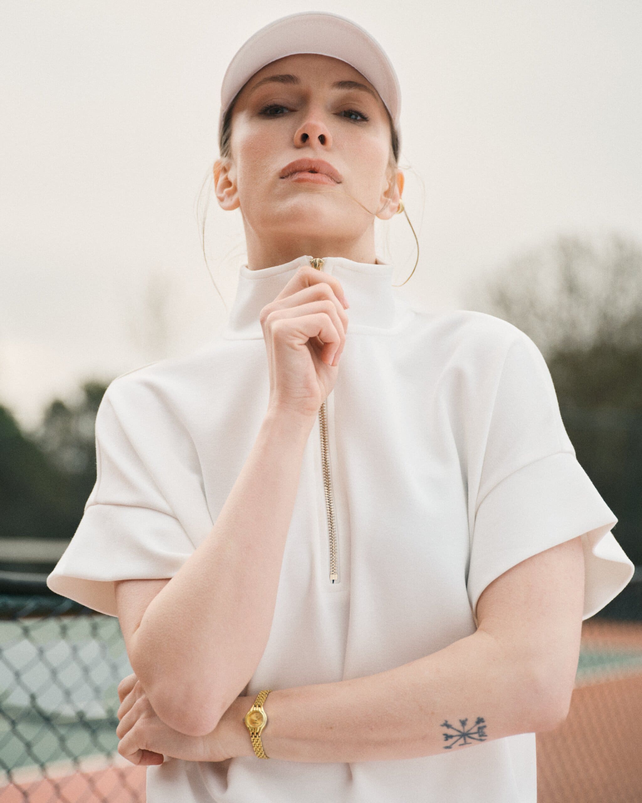 Low-angle portrait of Yasemin Kay Allen in a white visor and half-zip top, hand on the gold zipper, gold watch catching soft overcast light for LC Waikiki.