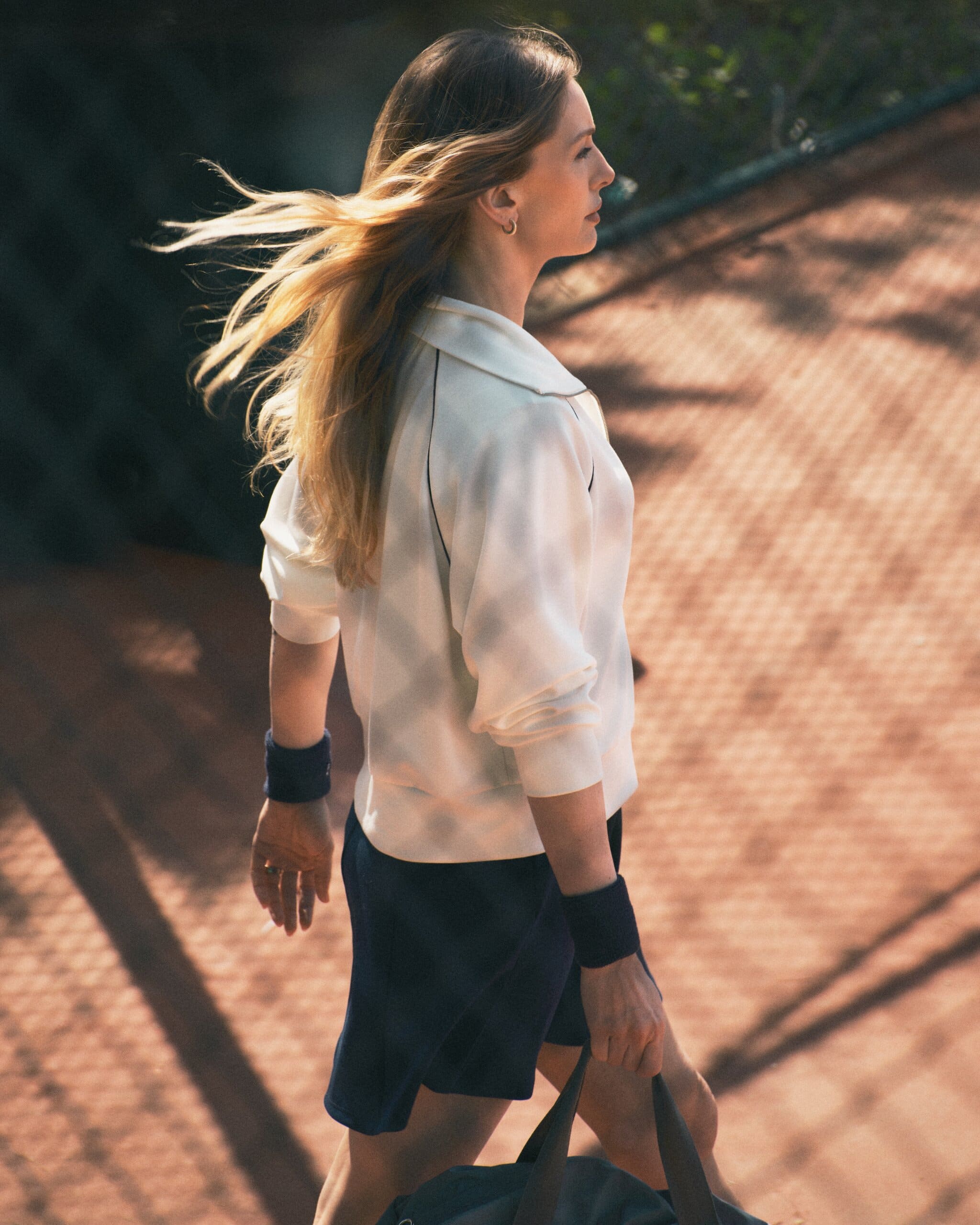 Yasemin Kay Allen strides across sunlit clay in profile, hair caught in the wind, navy wristbands against a white LC Waikiki track top.