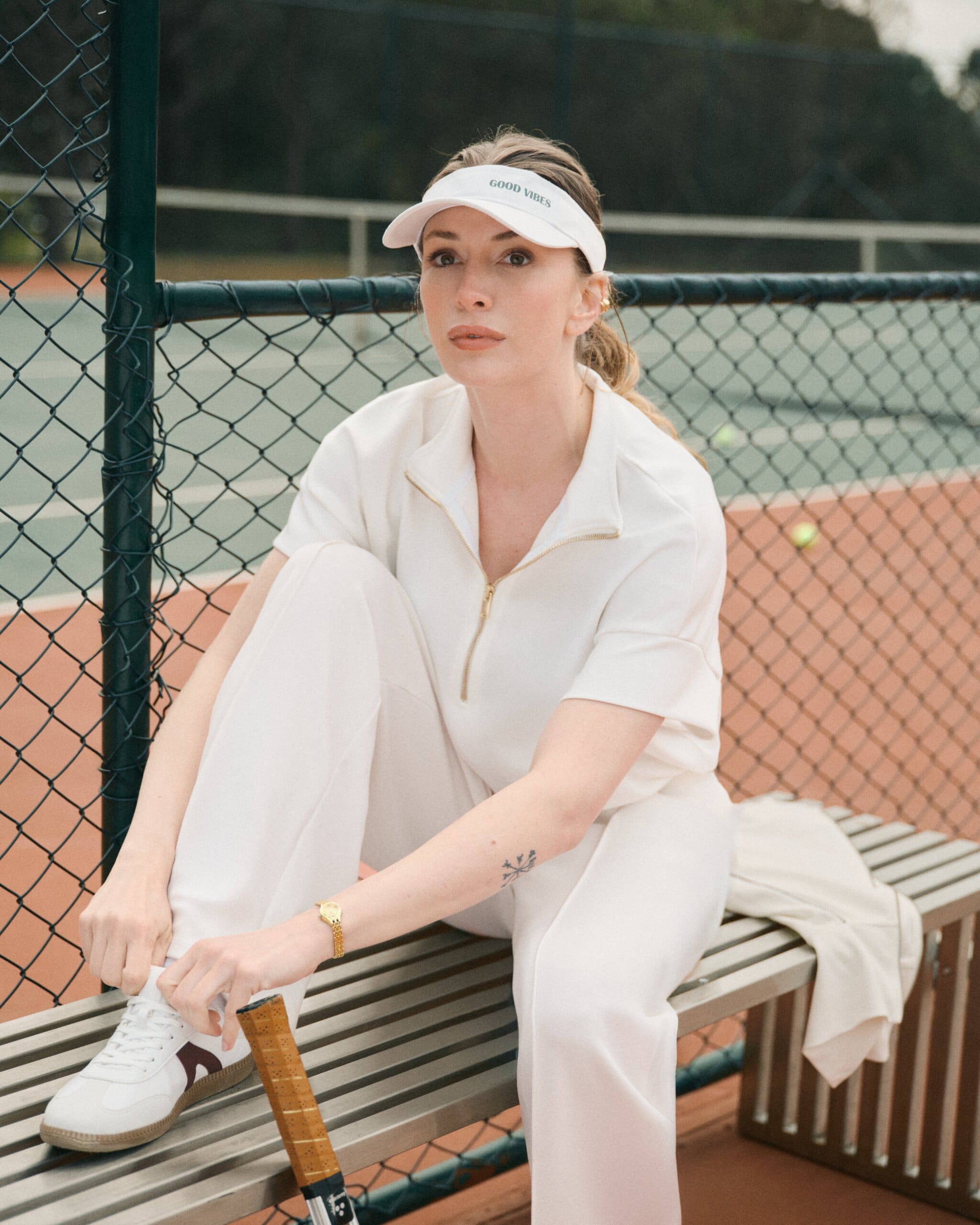 Yasemin Kay Allen sits on a courtside bench in head-to-toe cream with a Good Vibes visor, one hand at her sneaker and a racket propped beside her for LC Waikiki.