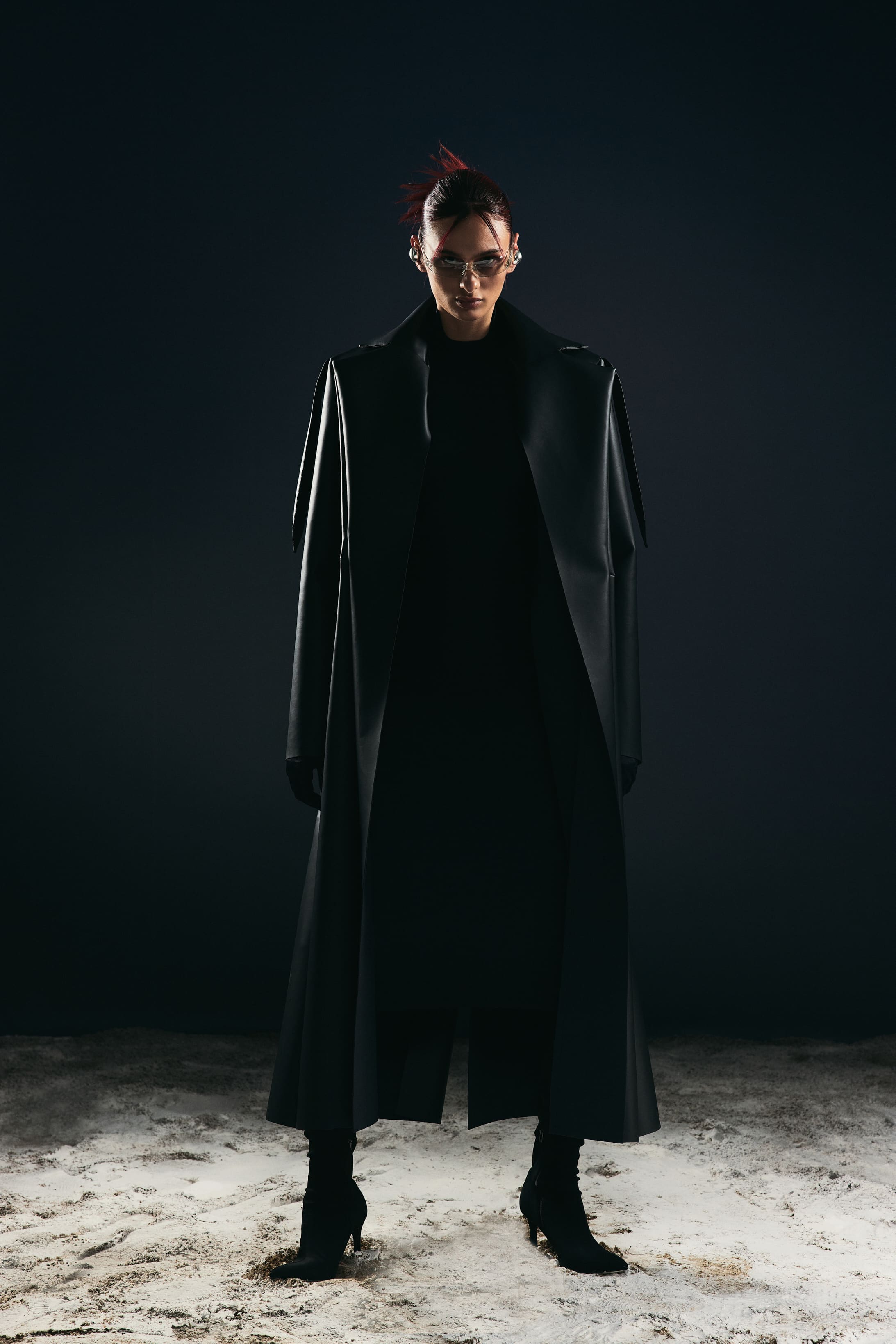 A matte black trench sweeps floor-length over pale sand, setting the shadowed tone of the Mysamoon campaign.