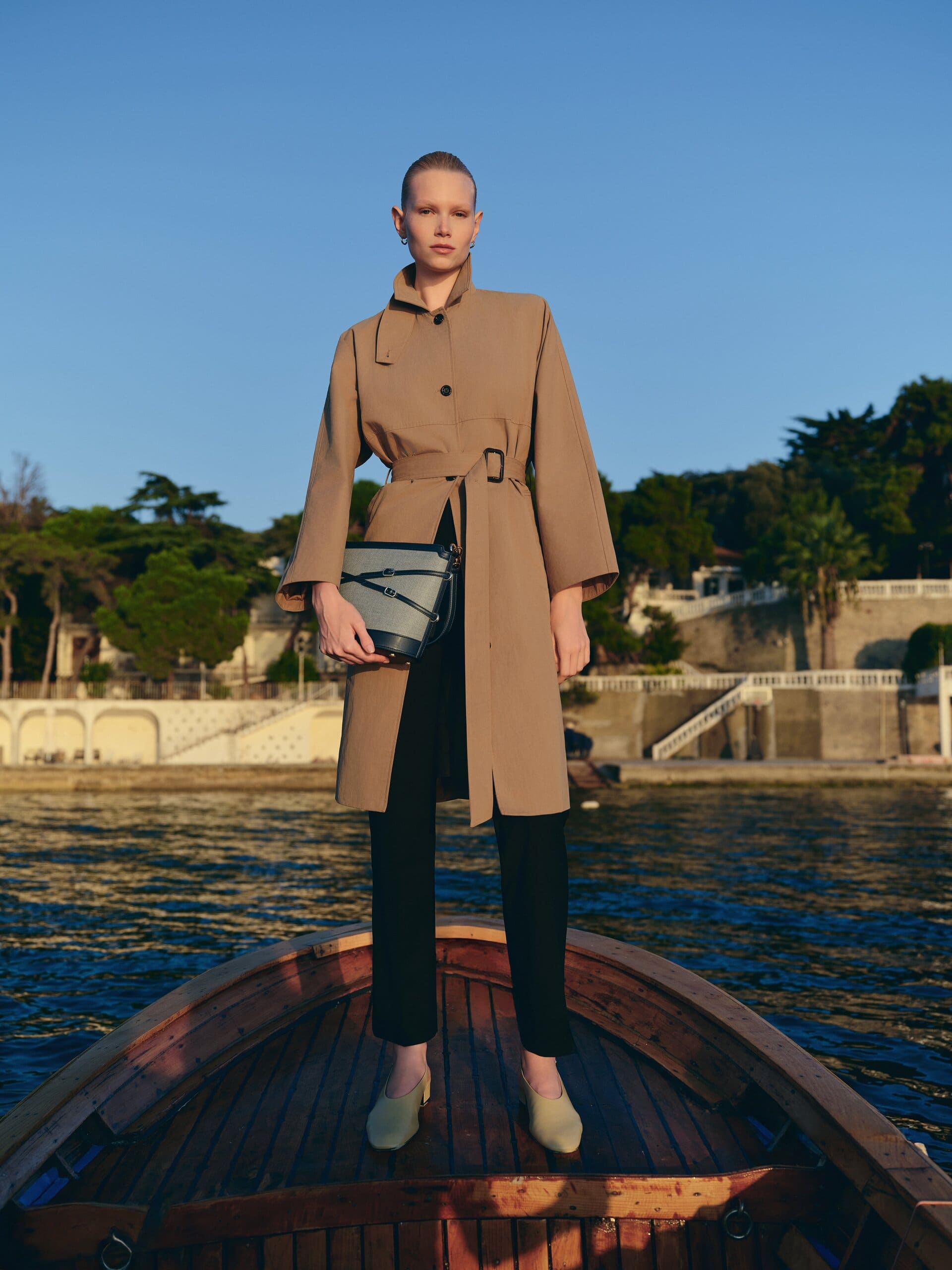 Standing square on the prow of the wooden boat in a belted camel trench, the two-tone bucket bag held at the waist for Owlish Studio.
