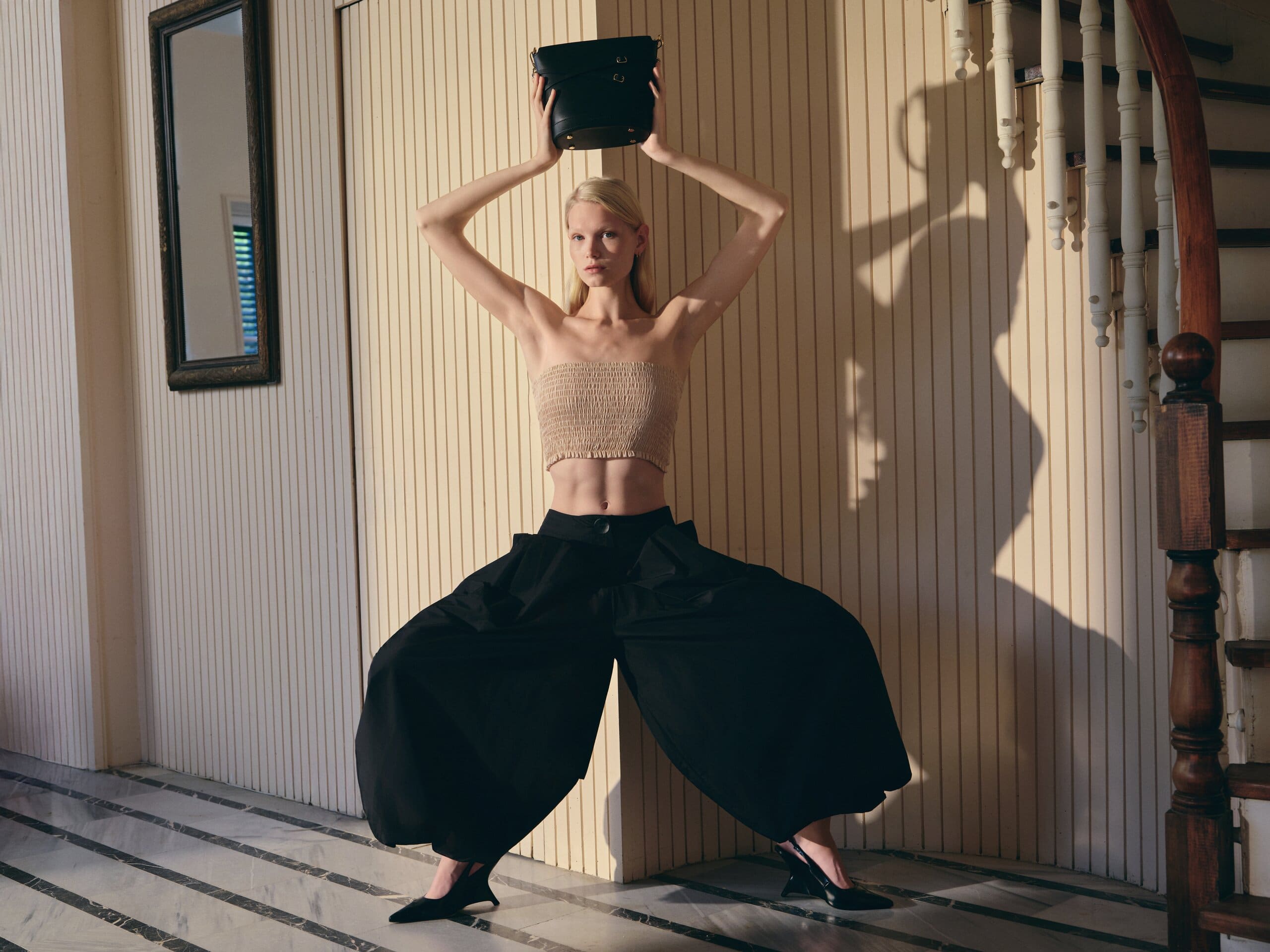 Wide stance in black tailoring with the bag raised overhead, hard noon shadows raking the cream slatted wall for Owlish Studio.