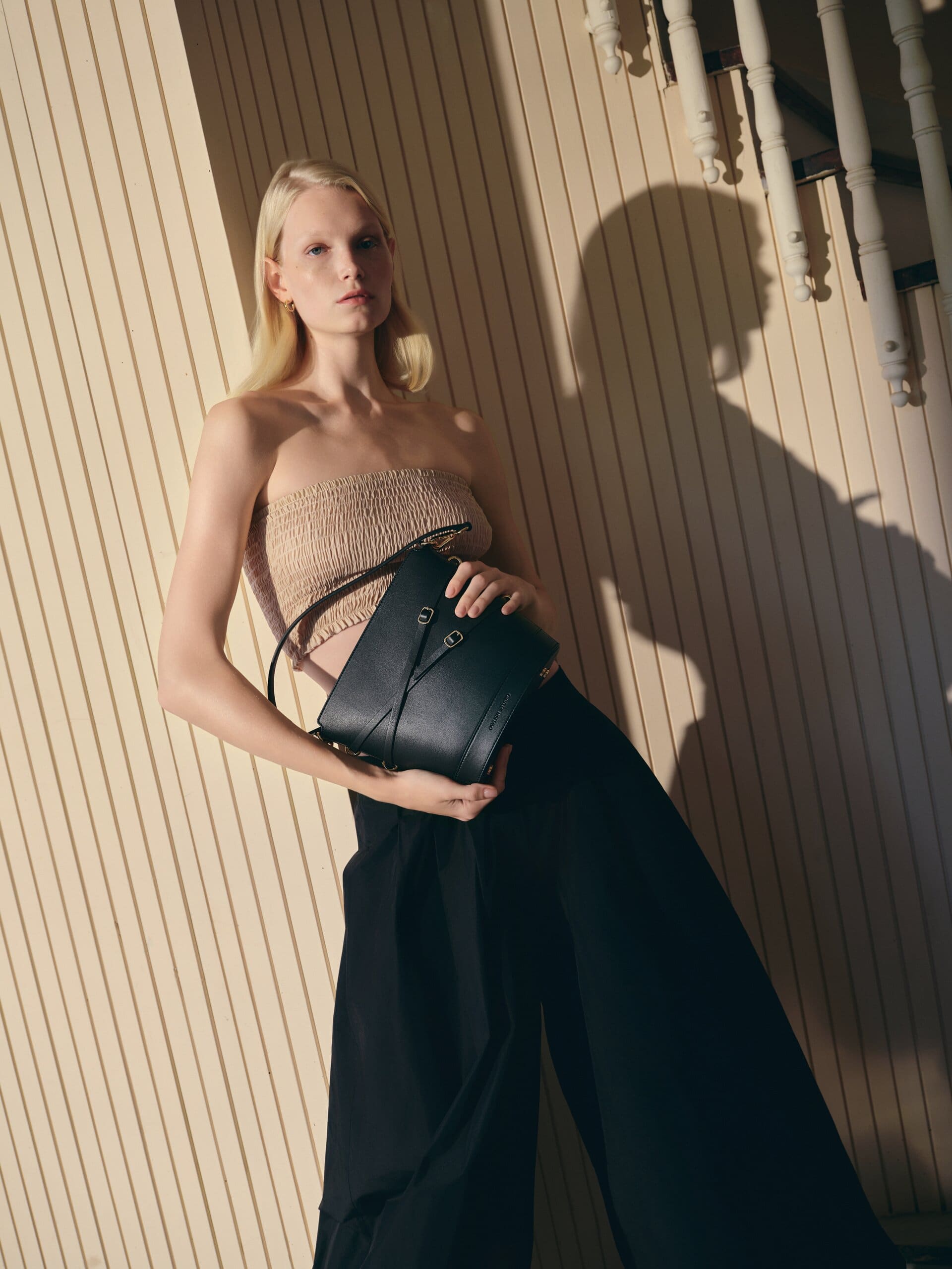 A nude smocked bandeau against the cream slatted wall, the black bucket bag held at the waist under a hard afternoon shadow for Owlish Studio.