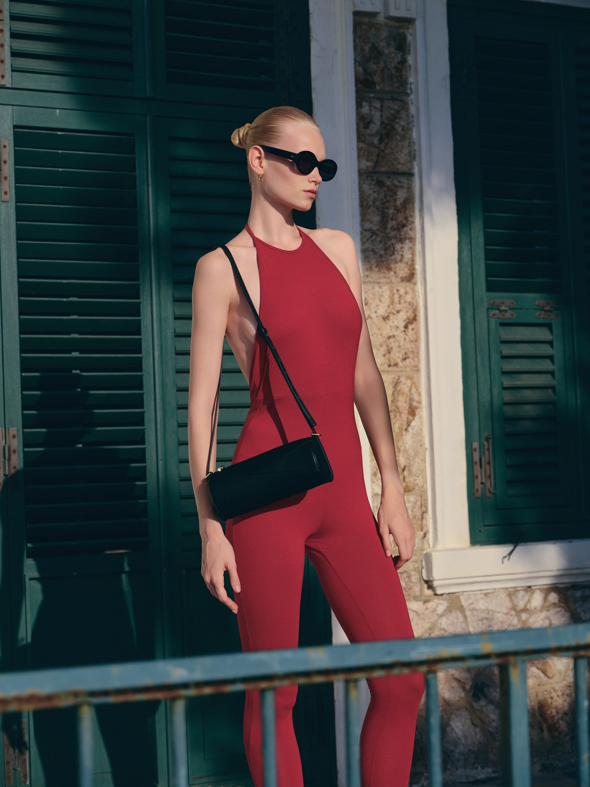 Wine-red halter catsuit and oval sunglasses against weathered green shutters, the black barrel bag slung across the body for Owlish Studio.