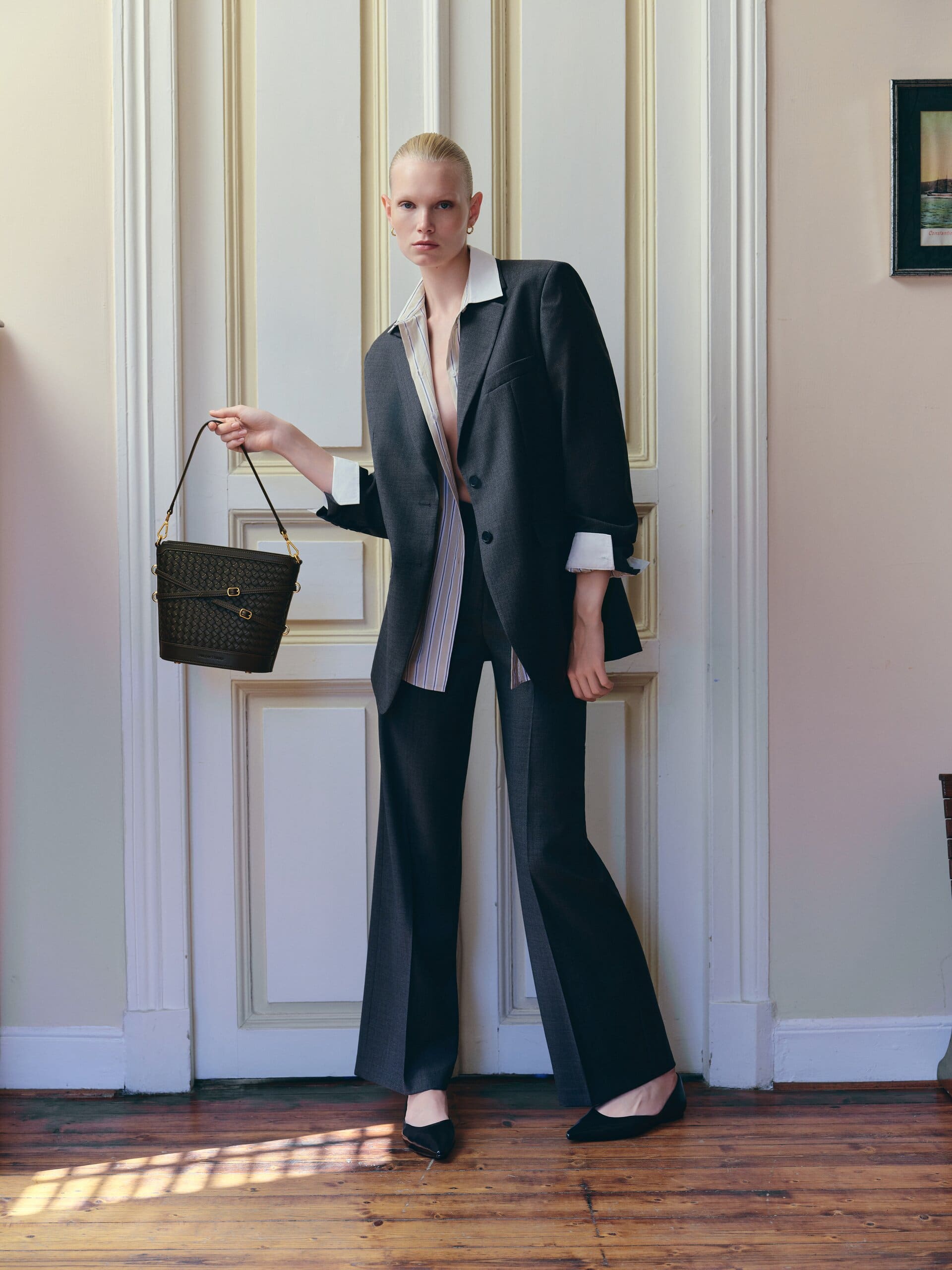 Full-length in the empty salon, the woven bucket bag held low as light rakes the old parquet for Owlish Studio.