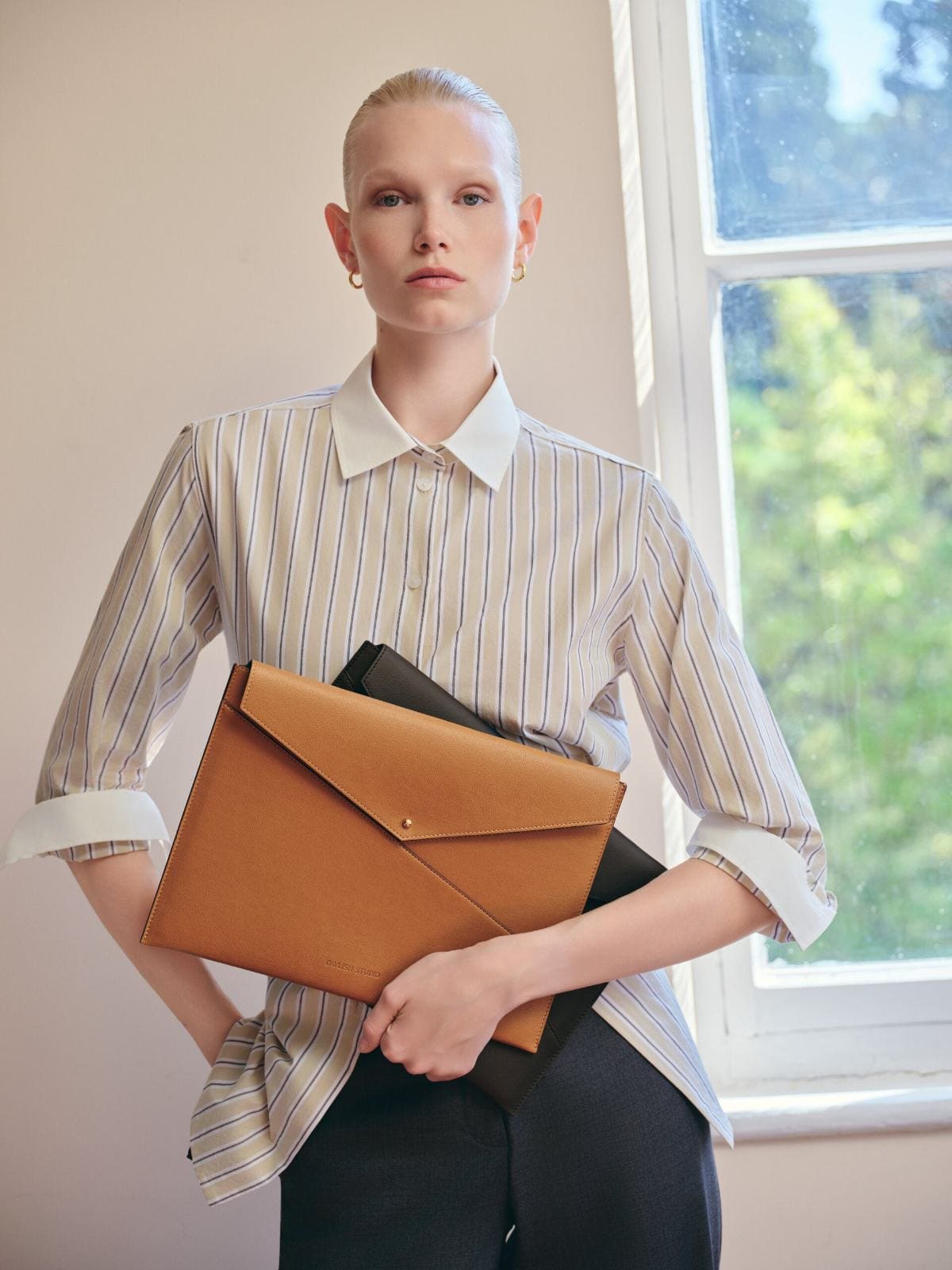 Striped poplin shirt and a camel envelope clutch held beside the folk-painted window for Owlish Studio.