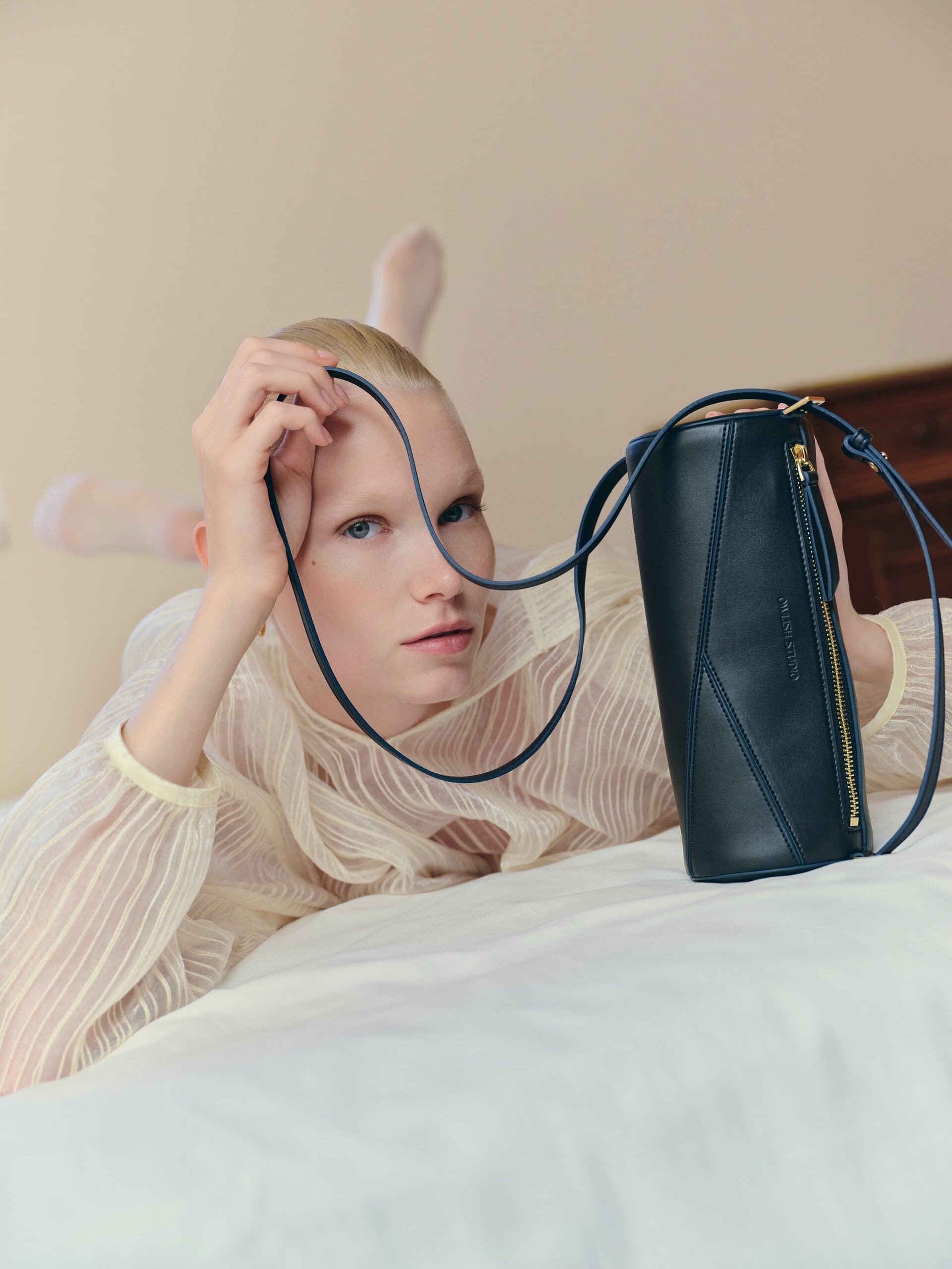 The navy pouch stood on end while its long strap loops through the fingers on the bed for Owlish Studio.