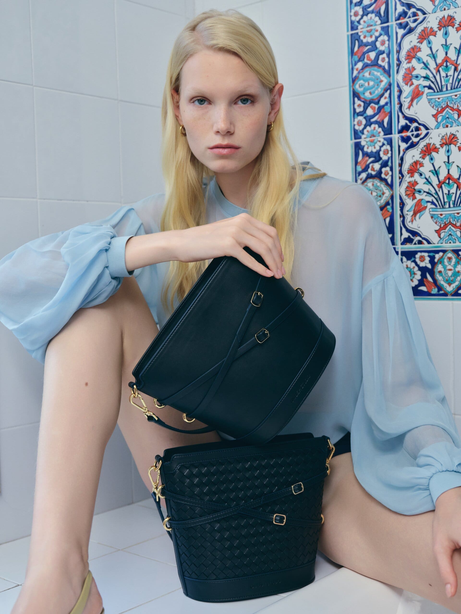 Seated in an Iznik-tiled hammam niche in a sheer indigo blouse, two navy bags stacked on the knee for Owlish Studio.