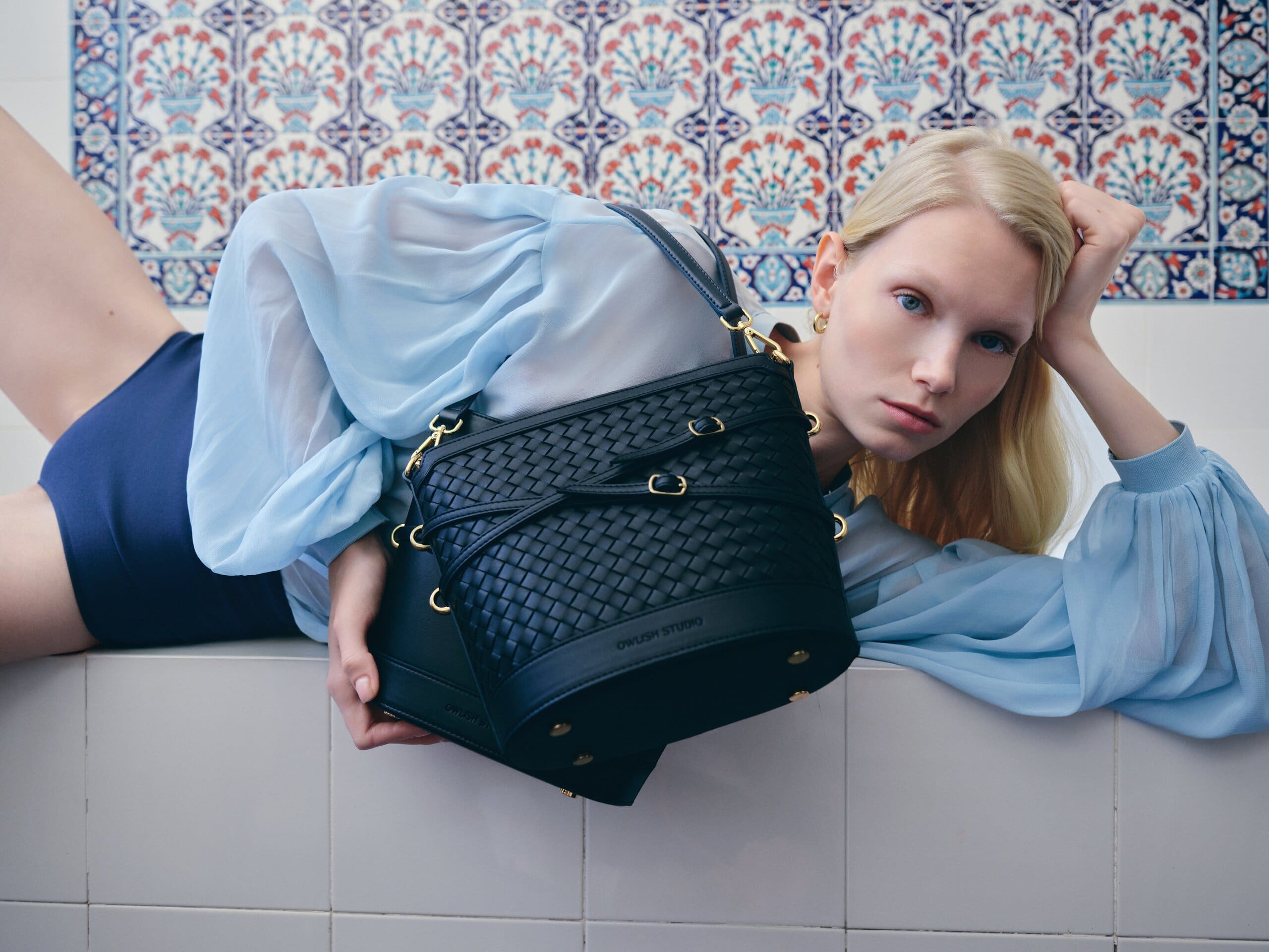 Tighter on the same ledge, the woven navy bucket bag filling the frame against blue and red tilework for Owlish Studio.